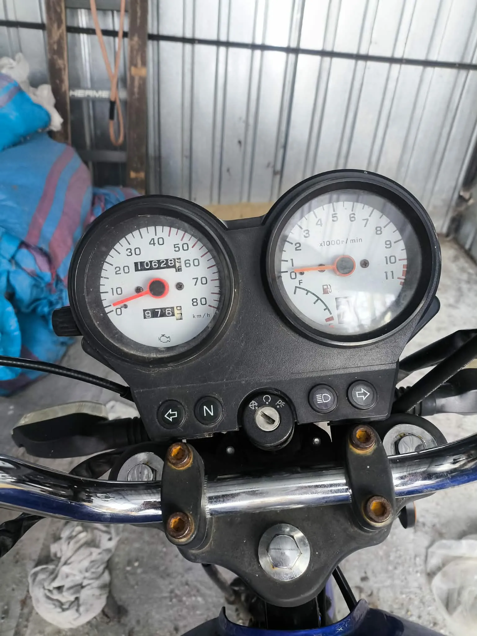 Zbliżenie na deski rozdzielcze motocykla z dwoma okrągłymi wskaźnikami. Po lewej stronie prędkościomierz z zakresem 0-80 km/h, wskazówką na 0, licznikiem przebiegu 10628 i dodatkowym wyświetlaczem 976. Po prawej tachometr z zakresem 0-11 x1000r/min, wskazówką na 0 oraz wskaźnikiem paliwa. Poniżej znajduje się czarna konsola z przyciskami: strzałką w lewo, literą N, kluczykiem, symbolem światła i strzałką w prawo. W tle metalowa ściana, niebiesko-fioletowa tkanina oraz betonowa podłoga. Widoczne są również czarne kierownice z metalowymi elementami.