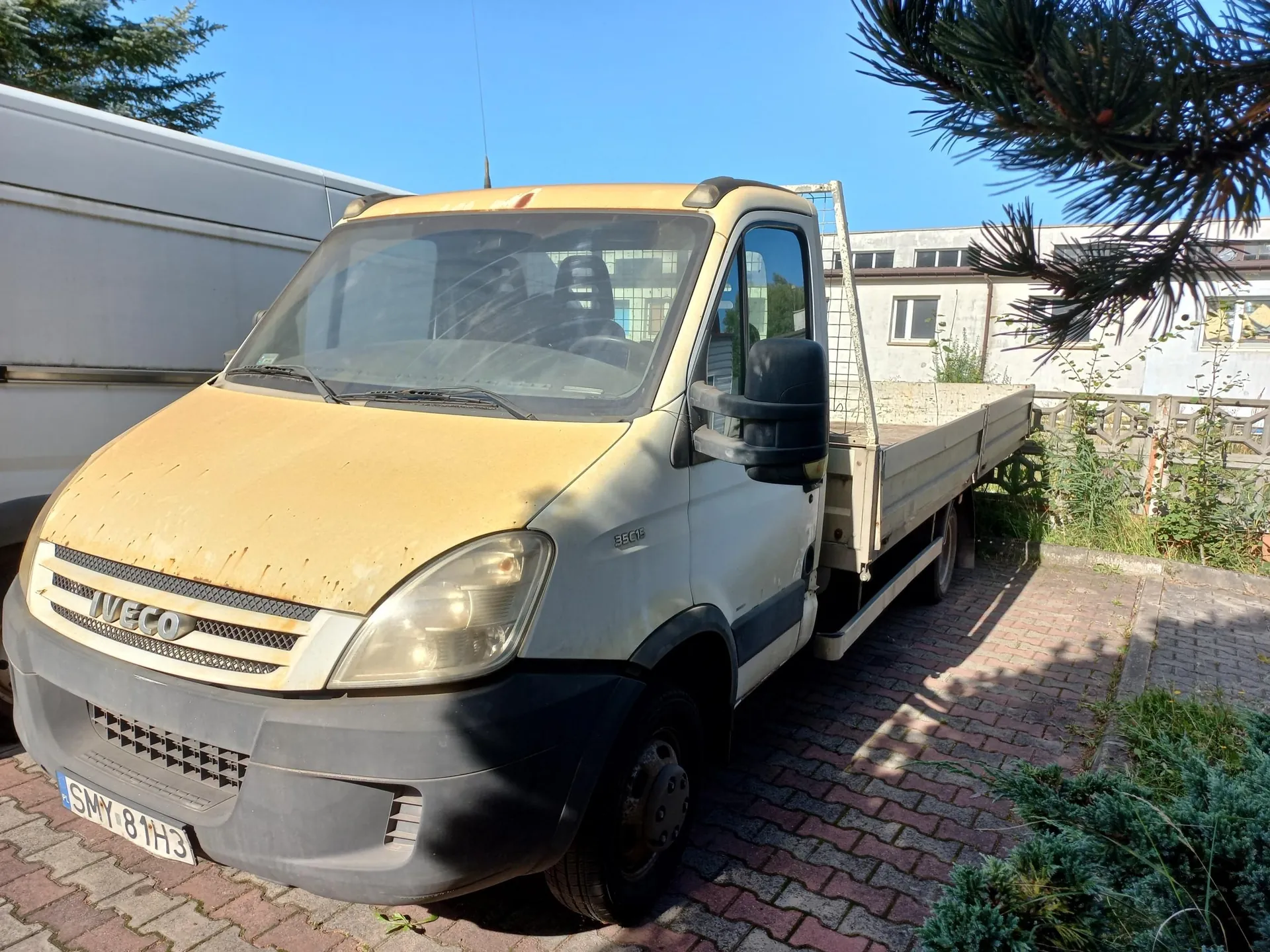 Żółty samochód dostawczy marki Iveco z otwartą platformą ładunkową, zaparkowany na kostce brukowej. Pojazd ma widoczne ślady zużycia i zabrudzenia na masce. Na przednim zderzaku znajduje się tablica rejestracyjna o numerze SMY 81H3. W tle widoczny jest biały furgon oraz jasny budynek.