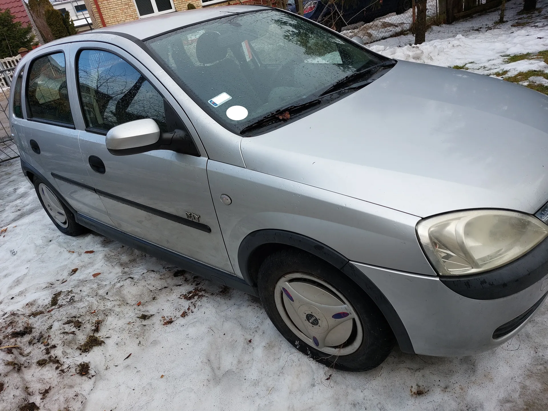 Srebrny samochód osobowy typu hatchback zaparkowany na śnieżnej powierzchni z widocznymi plamami trawy. Na boku pojazdu znajduje się napis "WY". Przednia szyba posiada dwa naklejki: białą okrągłą i prostokątną z niebieskim elementem. W tle widoczny jest ceglany dom z oknami, ogrodzenie oraz drzewa. Na kołach samochodu znajdują się felgi z logo. Śnieg pokrywa część terenu, a na ziemi leżą suche liście.