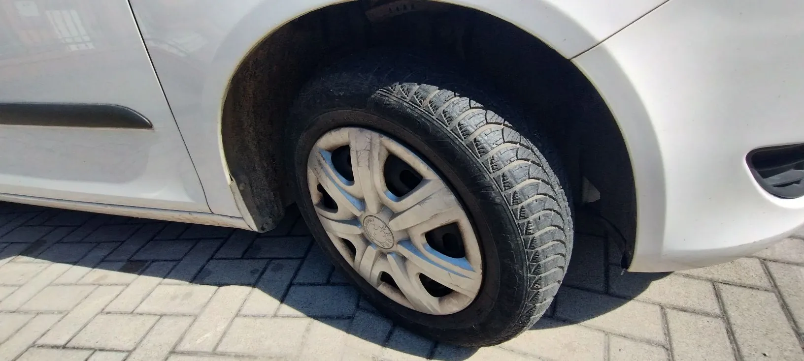 Zbliżenie na przednie prawe koło białego samochodu. Widoczna jest opona z wyraźnym bieżnikiem oraz srebrna felga z plastikowym kołpakiem. Samochód stoi na kostce brukowej.