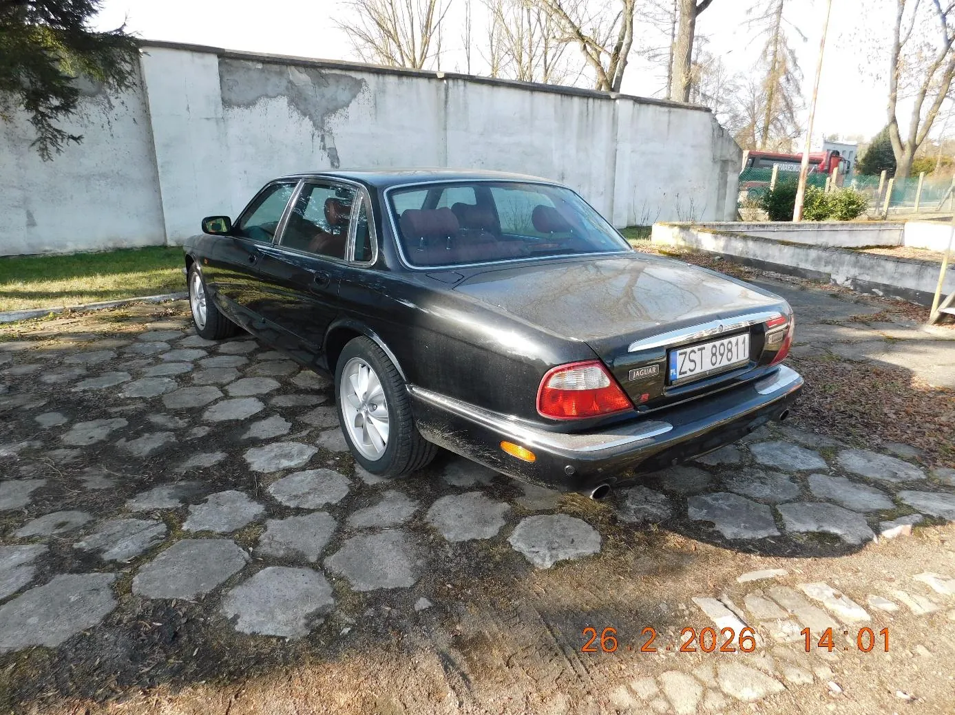 Czarny samochód marki Jaguar zaparkowany na brukowanej powierzchni. Widoczny tył pojazdu z tablicą rejestracyjną ZST 89811. W tle betonowa ściana, suche drzewa, trawa oraz pomarańczowy znaczek z datą 26.2.2026 i godziną 14:01 w prawym dolnym rogu. Na bagażniku widnieje napis JAGUAR.