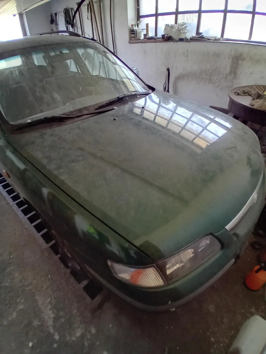 Zielony samochód pokryty kurzem znajduje się w garażu. W tle widać betonową podłogę, narzędzia wiszące na ścianie, okno z przedmiotami na parapecie oraz okrągły stół z ręcznikami. Na podłodze stoi pomarańczowy pojemnik.