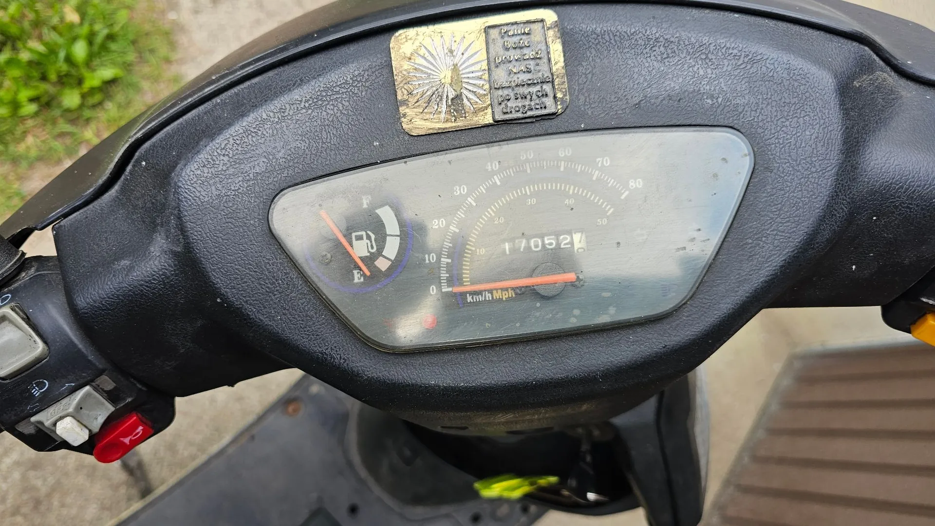 Zbliżenie na deskę rozdzielczą czarnego skutera. Widoczny jest analogowy prędkościomierz z podziałką w km/h i Mph, wskaźnik poziomu paliwa oraz licznik przebiegu wskazujący 17052 km. Nad zegarami znajduje się złota, ozdobna tabliczka z tekstem: 'Pamięć Wieczna przeżył 24 lata i 3 miesiące wędrując po swych drogach'.
