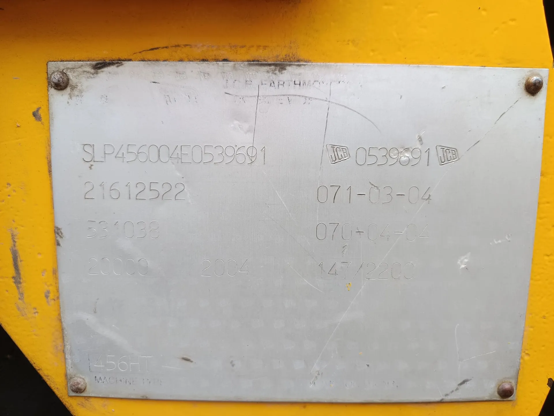 Srebrna tabliczka znamionowa przymocowana do żółtej powierzchni za pomocą czterech śrub. Na tabliczce znajdują się wyryte numery i kody: 'SLP456004E0539691', '21612522', '531038', '20000', '2004', '0539691' obok logo JCB, '071-03-04', '070-04-04', '147/2200' oraz w dolnej części '456HT' i napis 'MACHINE TYPE'.