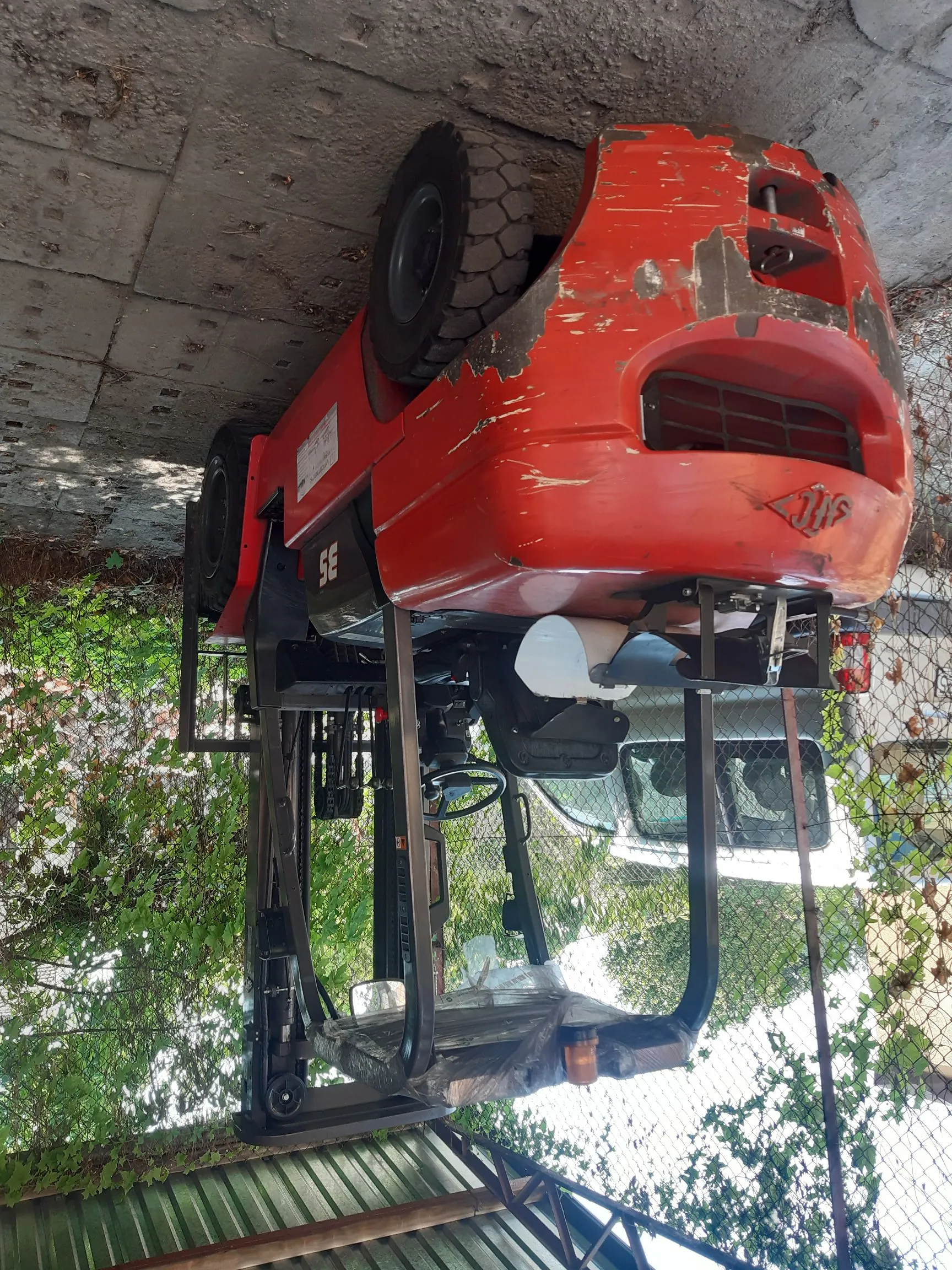 Czerwony wózek widłowy marki SE zaparkowany na betonowym podłożu obok metalowego ogrodzenia i zielonych drzew. Maszyna jest widoczna z boku i z tyłu, posiada widoczne ślady zużycia i odpryski farby na tylnej obudowie.