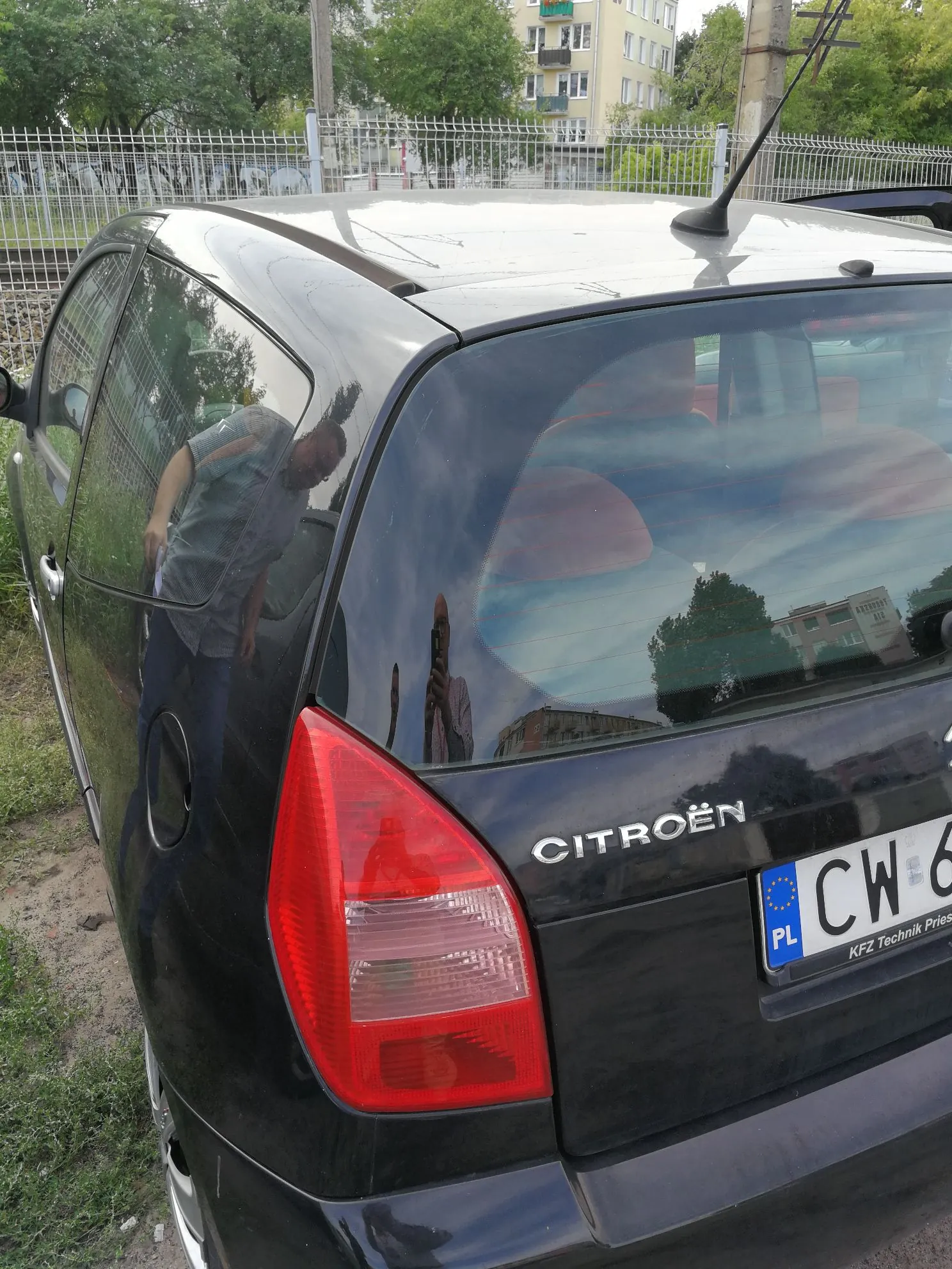 Tylna część czarnego samochodu marki Citroën. Na klapie bagażnika widnieje napis CITROËN oraz fragment tablicy rejestracyjnej zaczynającej się od liter CW. W szybie samochodu odbija się postać osoby robiącej zdjęcie oraz otoczenie z budynkami i drzewami. W tle widoczny jest metalowy płot oraz wielopiętrowy budynek mieszkalny.