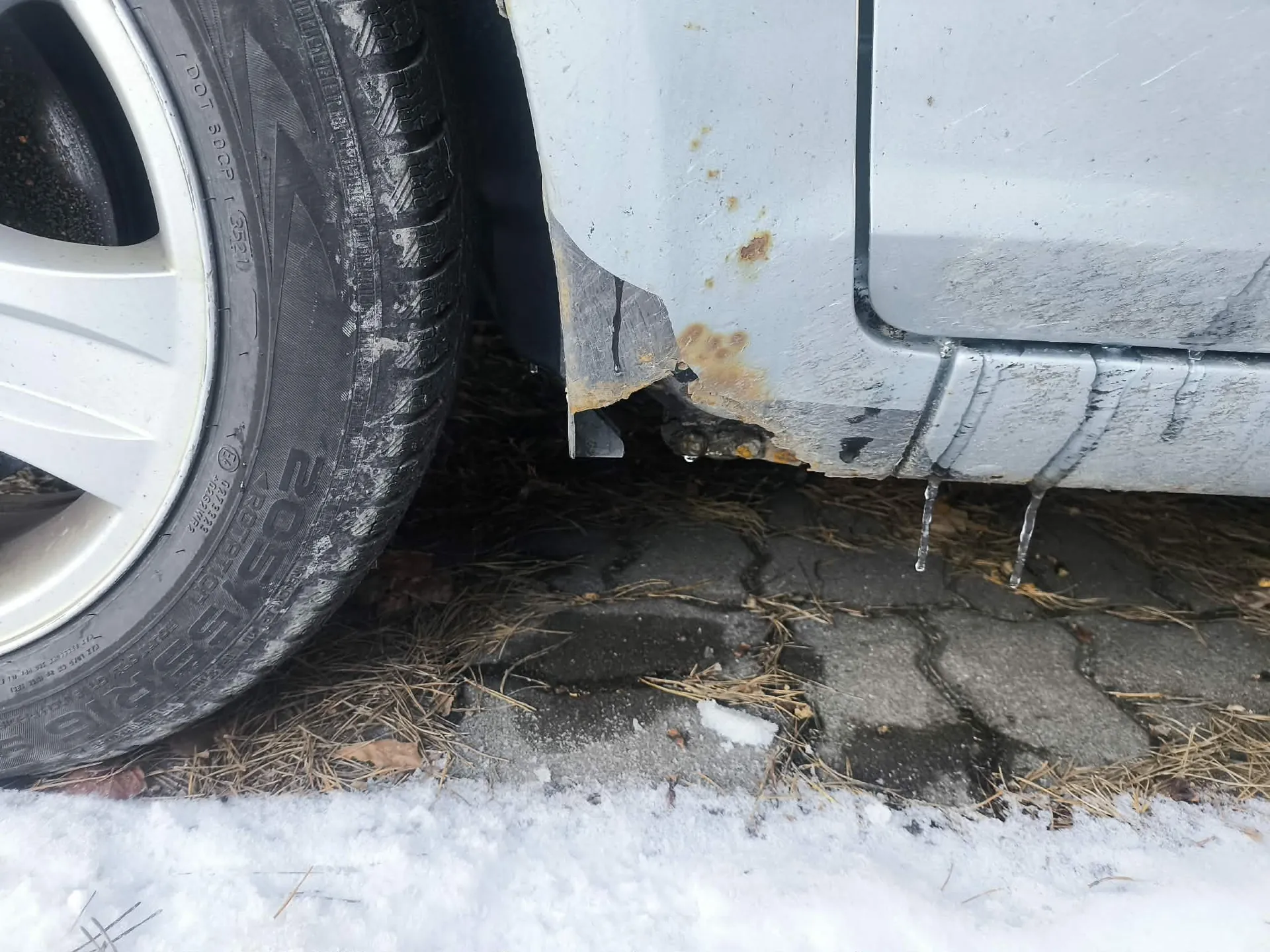 Zbliżenie na tylną część srebrnego samochodu z widocznym kołem o oznaczeniach 205/55 R16. Na karoserii widoczne są plamy rdzy oraz krople lodu zwisające z podwozia. Pod samochodem asfaltowa nawierzchnia z suchą trawą i śniegiem.