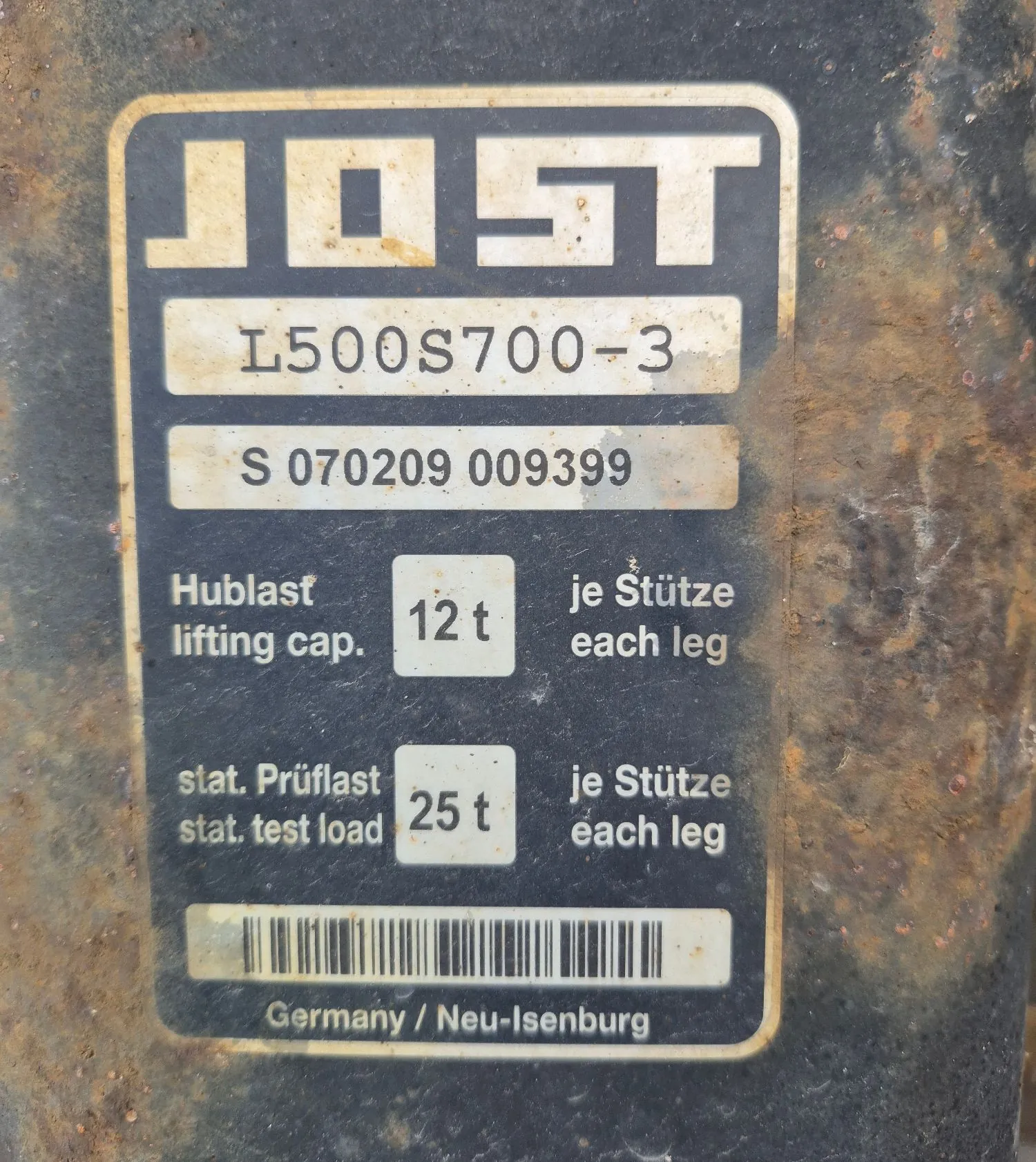 Zbliżenie na metalową tabliczkę znamionową firmy JOST. Na górze znajduje się duże logo 'JOST', poniżej model 'L500S700-3' oraz numer seryjny 'S 070209 009399'. Tabliczka zawiera dane techniczne w języku niemieckim i angielskim: 'Hublast / lifting cap. 12 t je Stütze / each leg' oraz 'stat. Prüflast / stat. test load 25 t je Stütze / each leg'. Na dole widnieje kod kreskowy oraz napis 'Germany / Neu-Isenburg'. Tabliczka jest przytwierdzona do zardzewiałej, ciemnej powierzchni metalowej.