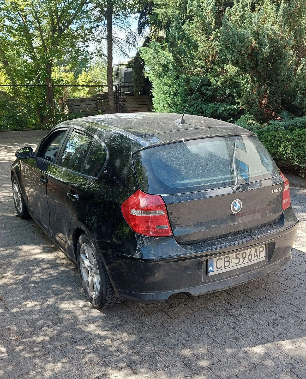 Czarny samochód marki BMW 118d zaparkowany na kostce brukowej. Pojazd jest widoczny od tyłu i z boku. Na klapie bagażnika znajduje się logo BMW oraz oznaczenie modelu 118d. Samochód ma polską tablicę rejestracyjną o numerze CB 596AP. W tle widoczne są zielone drzewa i krzewy.
