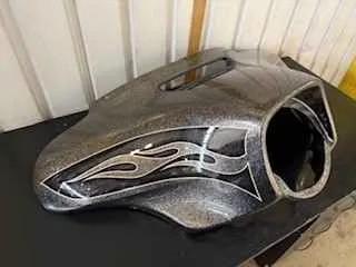 Metalowy zbiornik paliwa motocyklowy o srebrnym kolorze z czarnymi i białymi płomieniami, umieszczony na czarnej powierzchni. W tle białe pionowe panele ściany. Brak czytelnego tekstu.