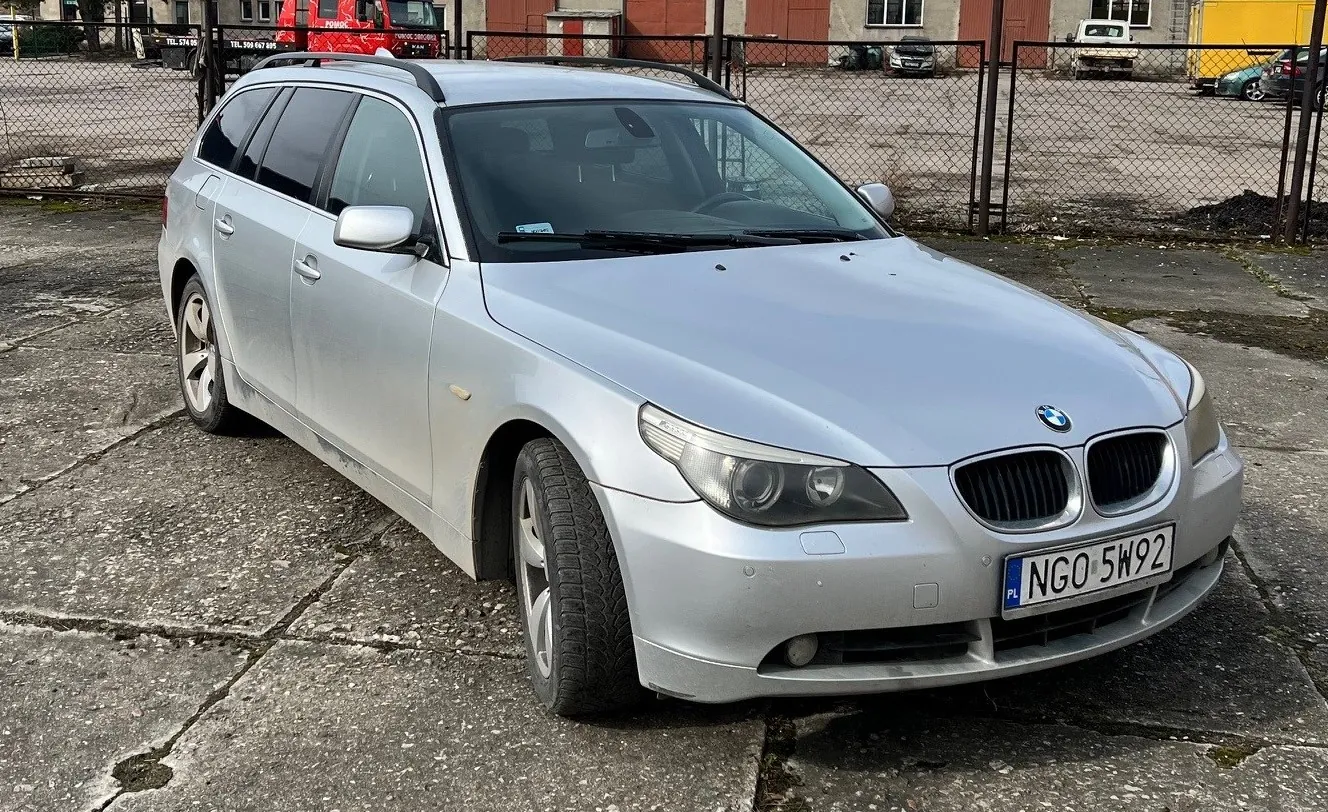 Srebrny samochód osobowy typu kombi marki BMW zaparkowany na pękniętym betonie. Widoczna tablica rejestracyjna z napisem PL NGO 5W92. W tle siatka ogrodzeniowa, przemysłowe budynki oraz inne pojazdy, w tym czerwony ciężarówka i biały samochód. Podłoga pokryta jest pęknięciami i plamami mchu.