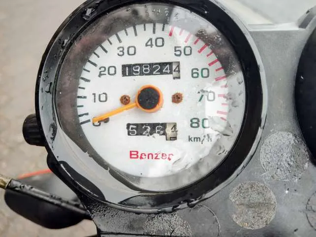 Zbliżenie na zegar prędkościowy motocykla lub skutera marki Benzer. Biała tarcza z czarnymi i czerwonymi oznaczeniami prędkości od 0 do 80 km/h. Czarny cyfrowy licznik przebiegu pokazuje 19824, a poniżej niego licznik podróży 5264. Pomarańczowa wskazówka prędkości wskazuje blisko 0 km/h. Na dole tarczy widnieje czerwony napis Benzer. Tarcza jest lekko zabrudzona, z widocznymi plamami i zadrapaniami. Czarna obudowa zegara oraz metalowa część z okrągłymi śladami zużycia po prawej stronie.
