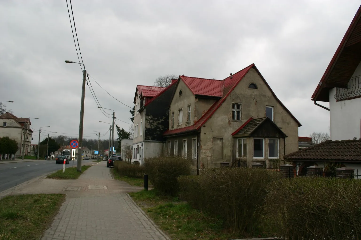 Zdjęcie przedstawiające dwupiętrowy dom z czerwonym dachem i szarymi, częściowo zniszczonymi tynkami na ścianach. Dom posiada dobudówkę z przeszklonymi oknami na parterze. Obok domu znajduje się chodnik z kostki brukowej, wzdłuż którego rosną niskie, przycięte krzewy. Po lewej stronie widoczna jest asfaltowa droga z samochodami, latarnie uliczne oraz znak drogowy zakazu wjazdu. W tle widać inne budynki mieszkalne pod zachmurzonym niebem.