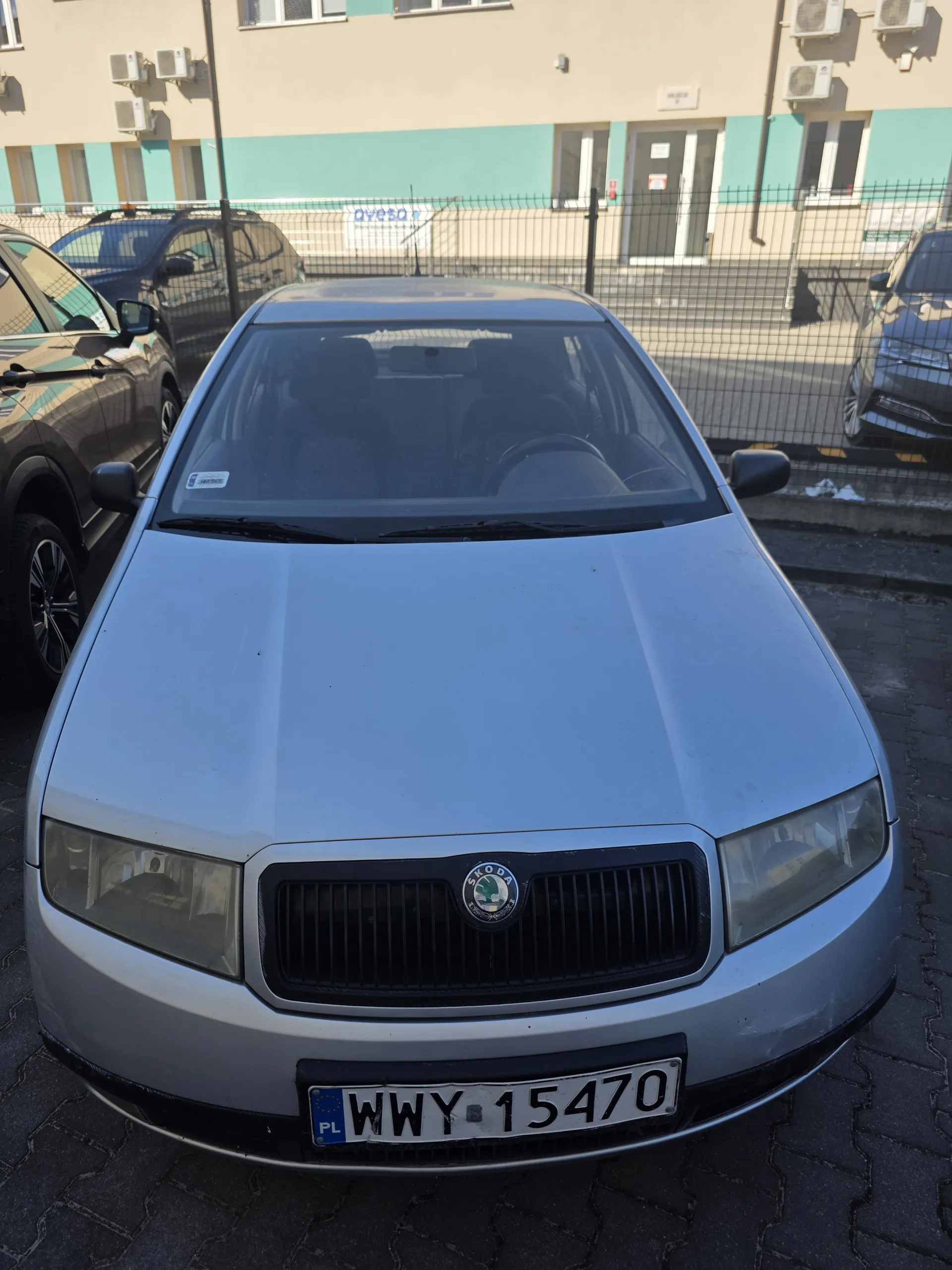 Srebrny samochód marki Skoda z tablicą rejestracyjną WWY 15470 (PL) zaparkowany na brukowanej powierzchni. W tle budynki z zielonymi i beżowymi elewacjami, klimatyzatory oraz inne zaparkowane pojazdy. Widoczne są siatki ochronne i fragmenty innych obiektów.
