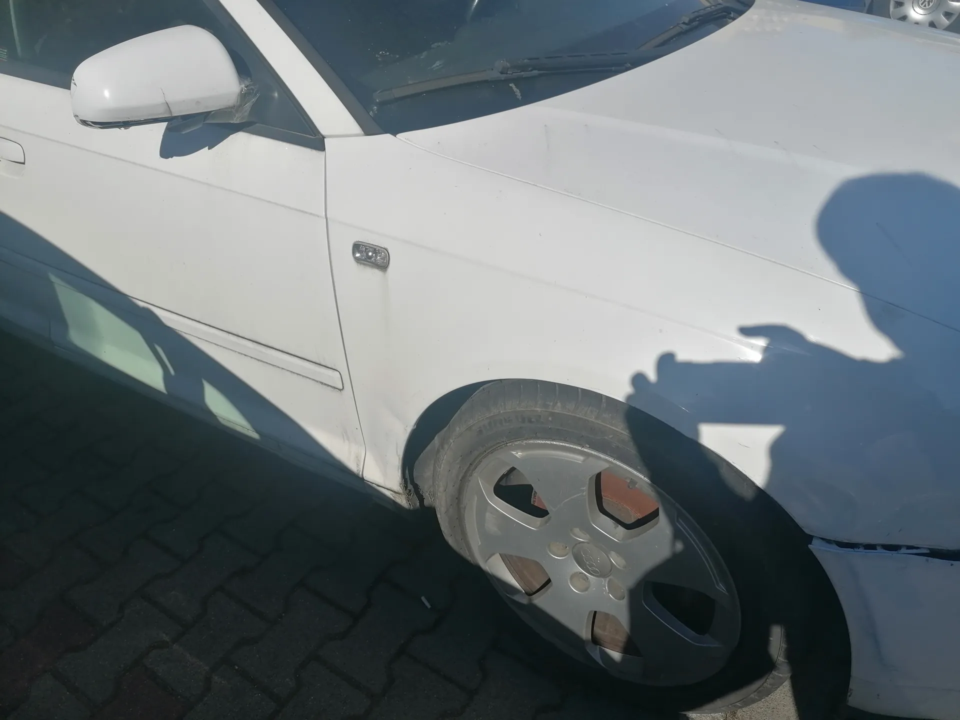 Biały samochód widziany z boku, z przodu, z widocznym lewym lusterkiem bocznym, przednim kołem z srebrną felgą z logo Audi, uszkodzeniem nad kółkiem oraz cieniem osoby trzymającej urządzenie fotografujące. Samochód stoi na brukowanej powierzchni. Widoczne są przednie wycieraczki i część przedniej szyby.