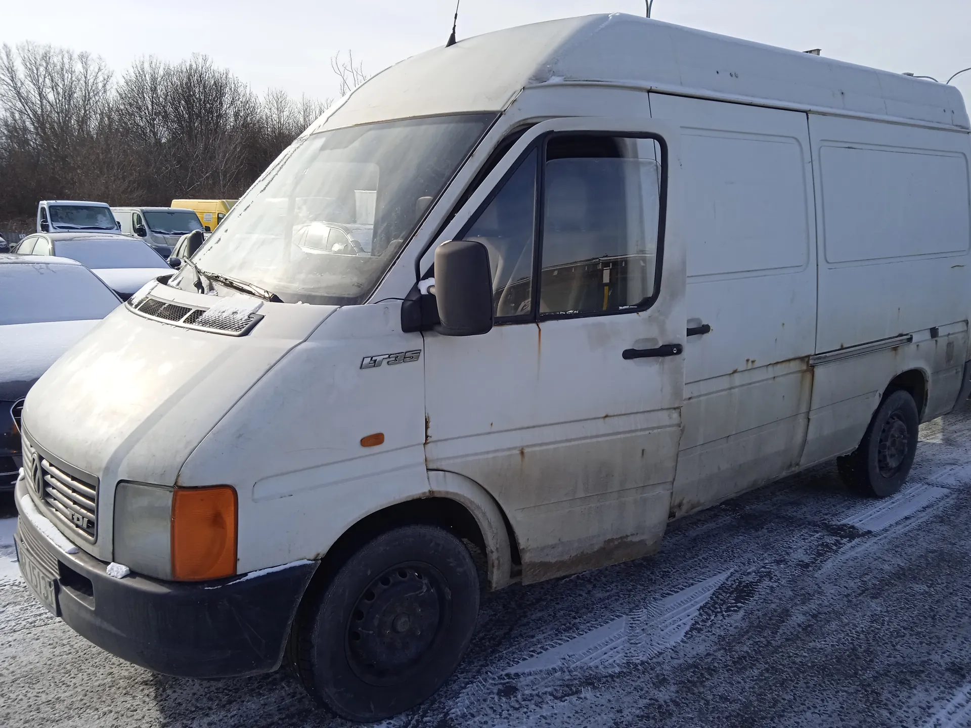 Biały furgon marki Volkswagen LT35 z widocznymi śladami rdzy na bokach i dolnej części nadwozia. Na masce oraz przedniej szybie leży niewielka ilość śniegu. Furgon stoi na zasypanej śniegiem nawierzchni, w tle widoczne są inne pojazdy oraz drzewa bez liści. Na boku pojazdu znajduje się napis "LT35". Oświetlenie wskazuje na zimowy dzień.