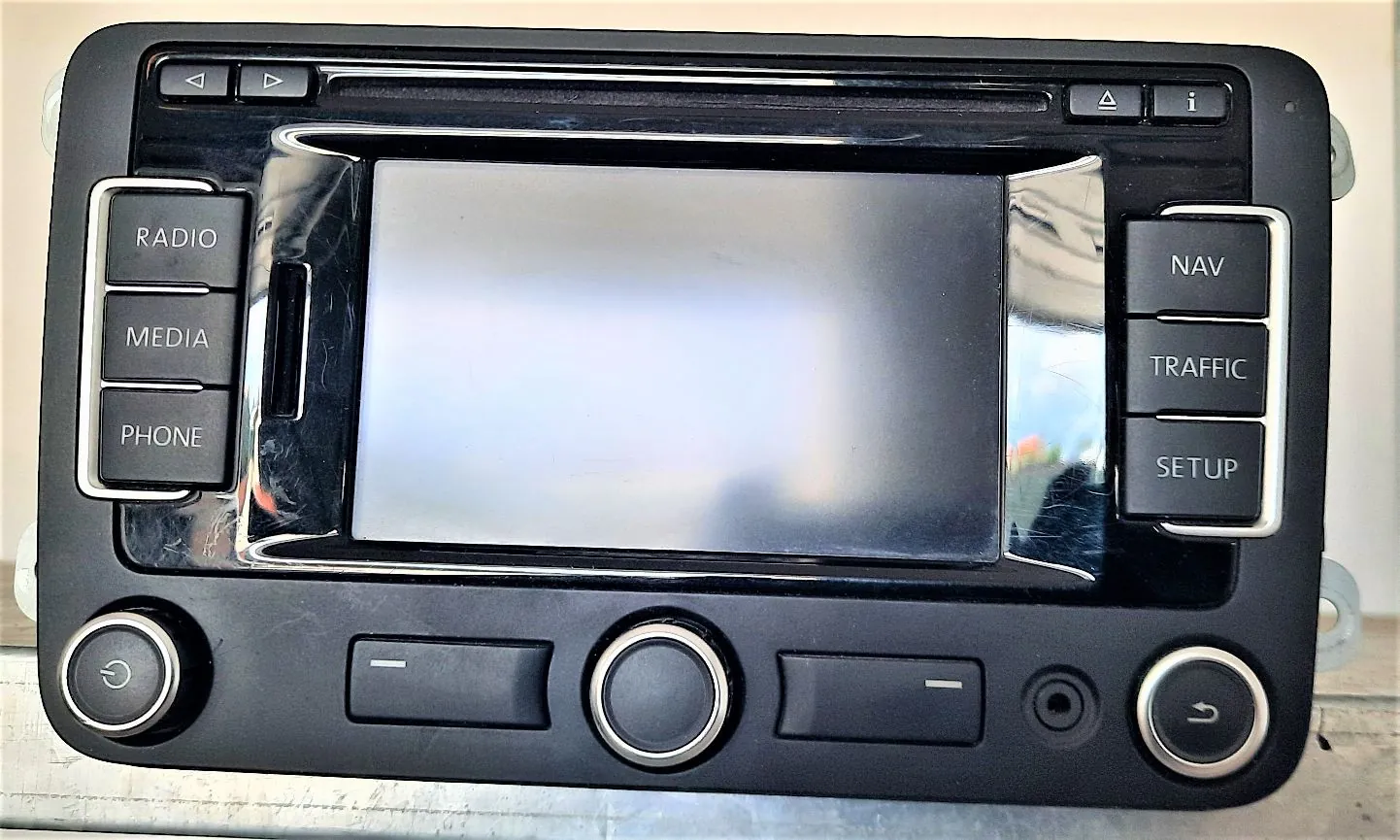 Czarny panel radia samochodowego z ekranem LCD w centrum. Po lewej stronie znajdują się przyciski: RADIO, MEDIA, PHONE. Po prawej stronie przyciski: NAV, TRAFFIC, SETUP. Na górze znajdują się małe przyciski sterujące, a na dole dwa pokrętła oraz dwa podłużne przyciski. Obudowa jest wykonana z czarnego plastiku z błyszczącymi elementami wokół ekranu.