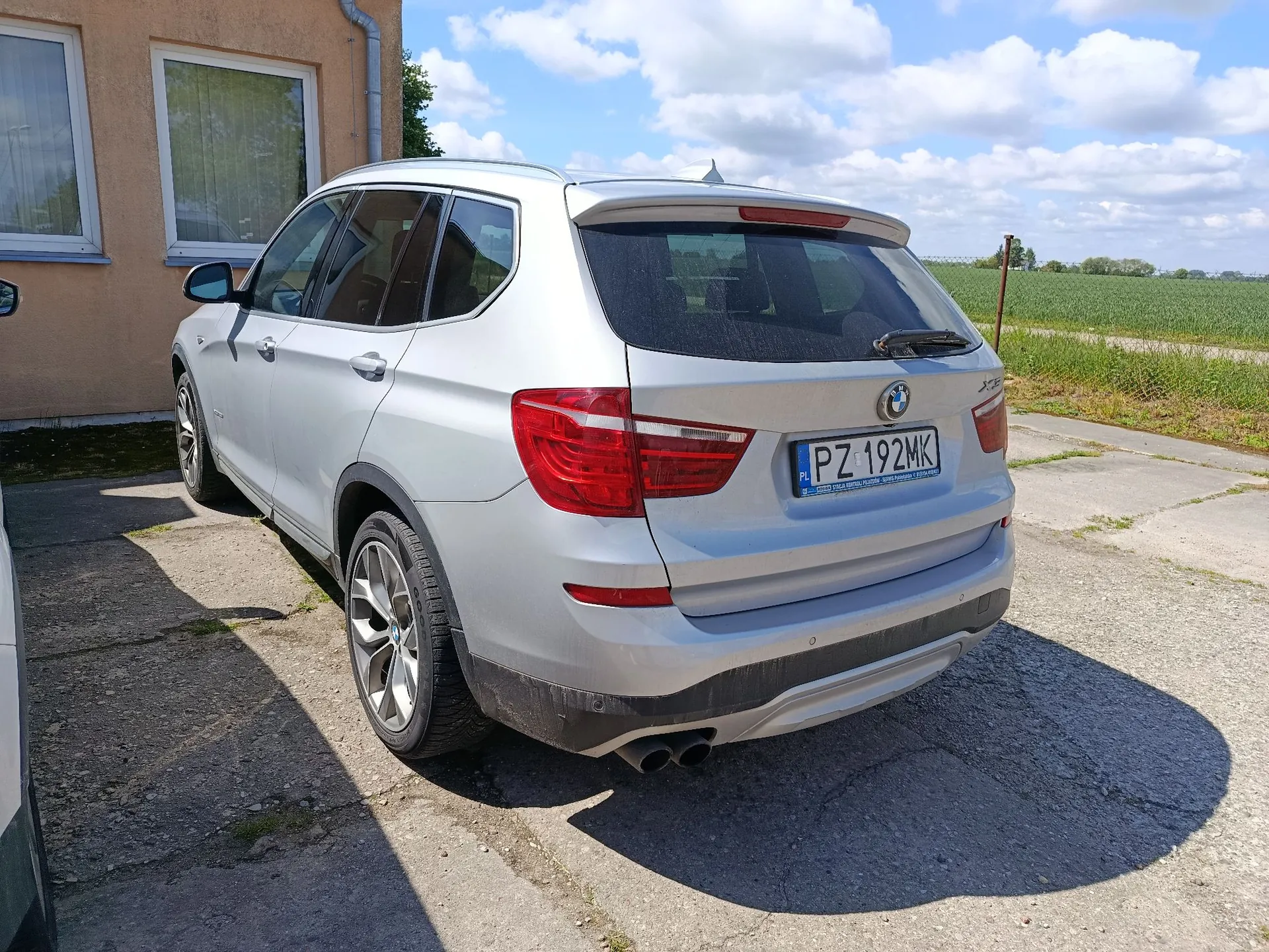 Srebrny samochód marki BMW X5 zaparkowany tyłem do obiektywu na betonowym podjeździe obok beżowego budynku. Samochód ma polską tablicę rejestracyjną o numerze PZ 192MK. W tle widoczne jest zielone pole pod jasnym niebem z białymi chmurami.