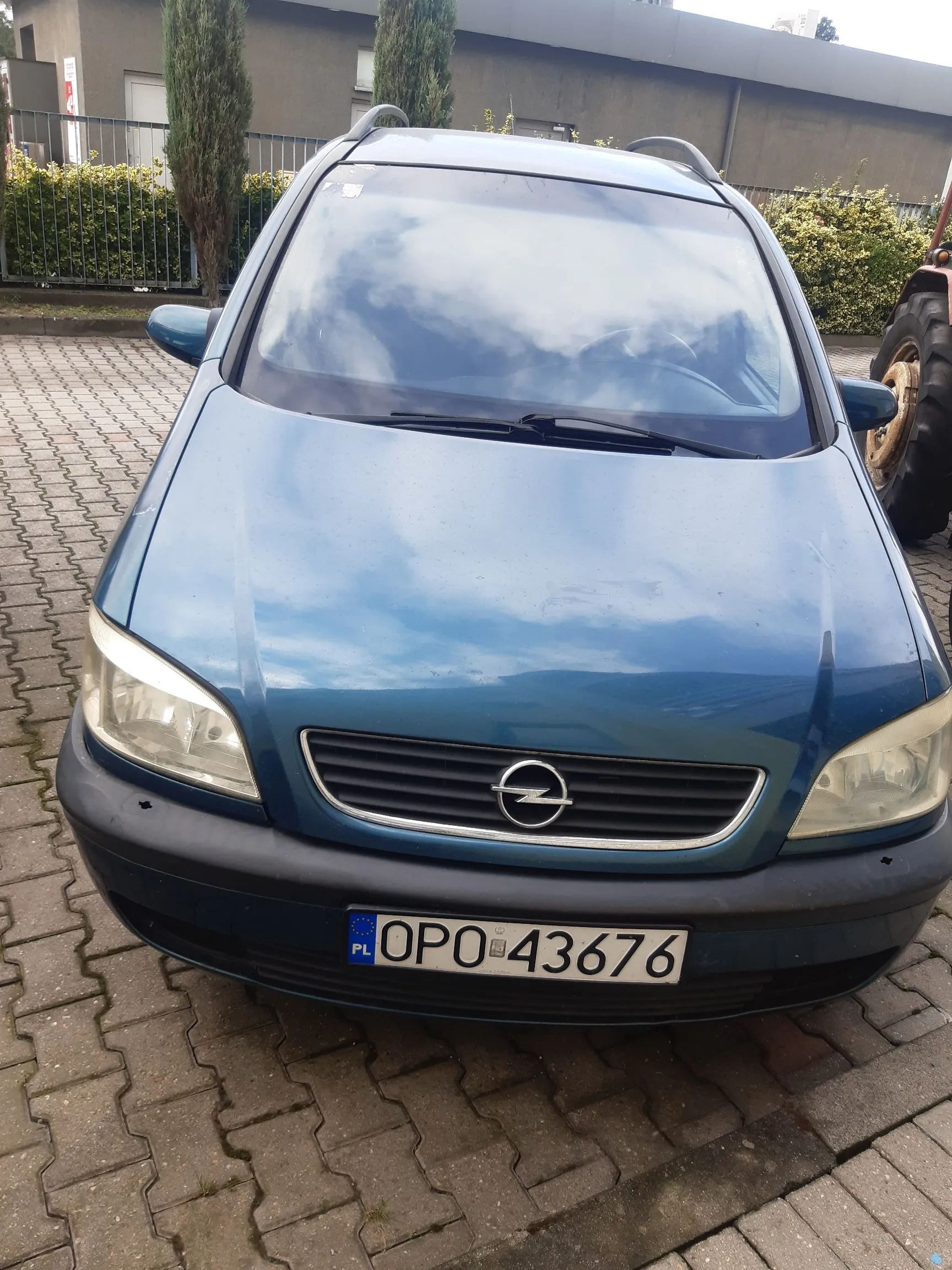 Niebieski samochód marki Opel zaparkowany na kostce brukowej. Widok z przodu, ukazujący maskę, przednie reflektory, grill z logo producenta oraz polską tablicę rejestracyjną o numerze OPO 43676. W tle widoczny jest budynek, zielone krzewy i fragment koła dużego pojazdu rolniczego po prawej stronie.