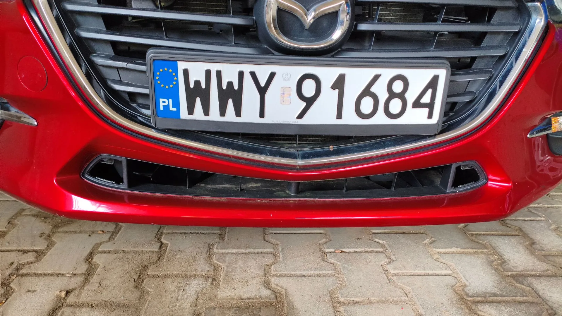 Zbliżenie na przednią część czerwonego samochodu marki Mazda. Widoczna jest polska tablica rejestracyjna o numerze WWY 91684 z symbolem Unii Europejskiej i oznaczeniem PL. Samochód stoi na kostce brukowej.