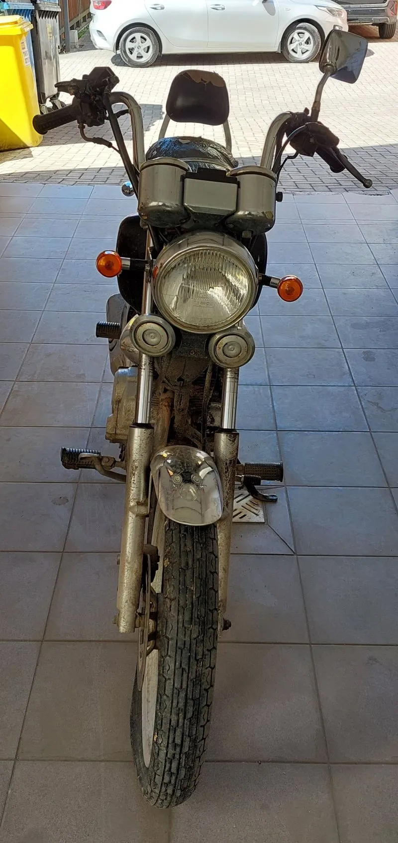 Przód motocykla z dużym reflektorem, pomarańczowymi kierunkowskazami i czarnym siedzeniem. Widoczne metalowe elementy z oznakami korozji. W tle biały samochód zaparkowany na brukowanej powierzchni oraz żółty pojemnik na śmieci. Motocykl stoi na szarych płytkach.