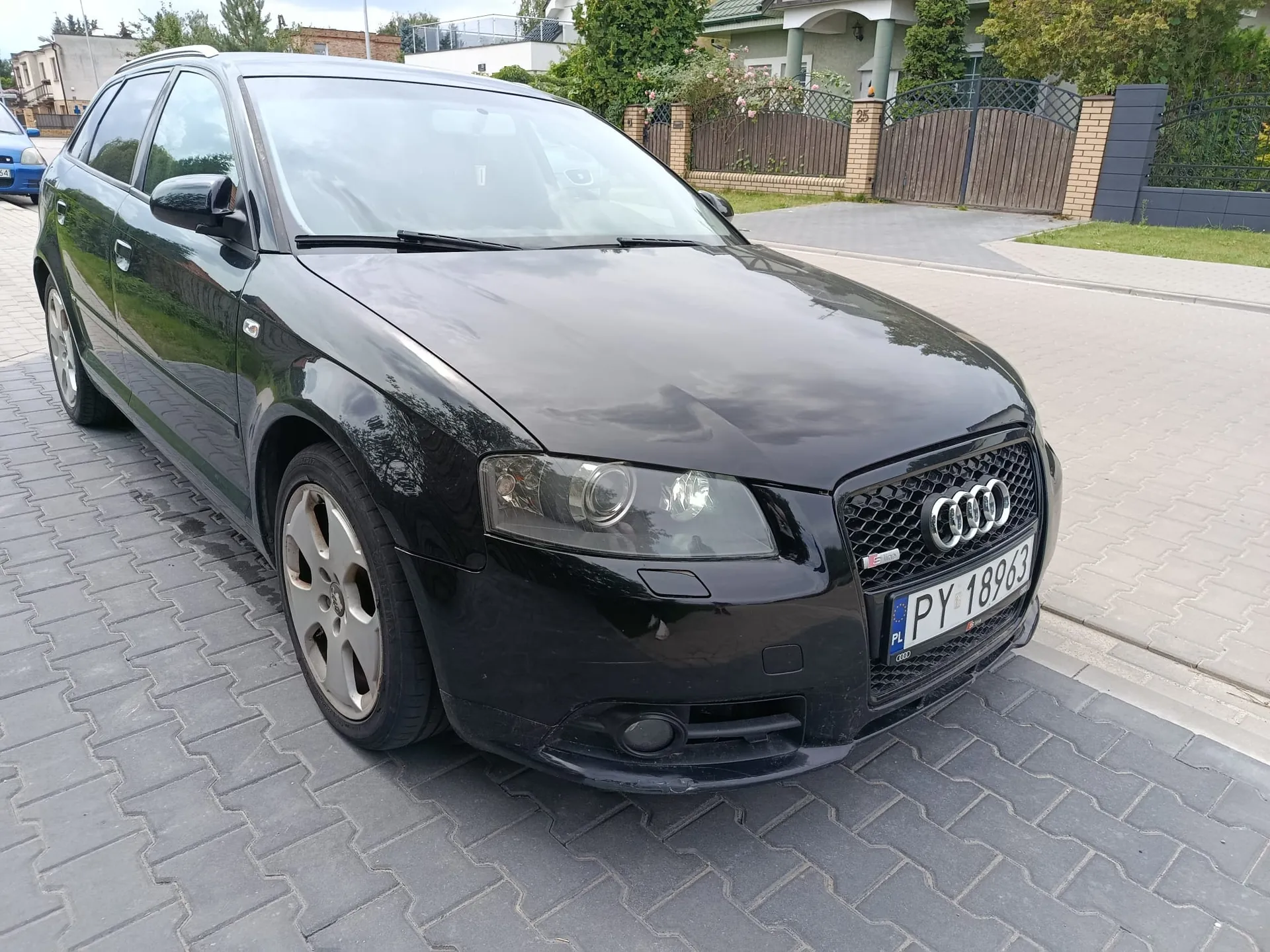 Czarny samochód marki Audi w nadwoziu kombi, zaparkowany na szarej kostce brukowej. Widoczny jest przód i lewy bok pojazdu. Samochód ma polską tablicę rejestracyjną o numerze PY 18963. W tle znajduje się dom z ogrodzeniem i zielenią.