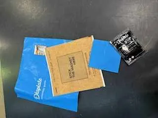 Na ciemnej powierzchni leżą: niebieska koperta z białym napisem "DHL", brązowy kartonik z napisami "THIS SIDE UP" i "FRAGILE", niebieski arkusz papieru oraz mały czarny pakiet z białym tekstem. Obiekty są ułożone na płasko, bez widocznych osób lub dodatkowych elementów.