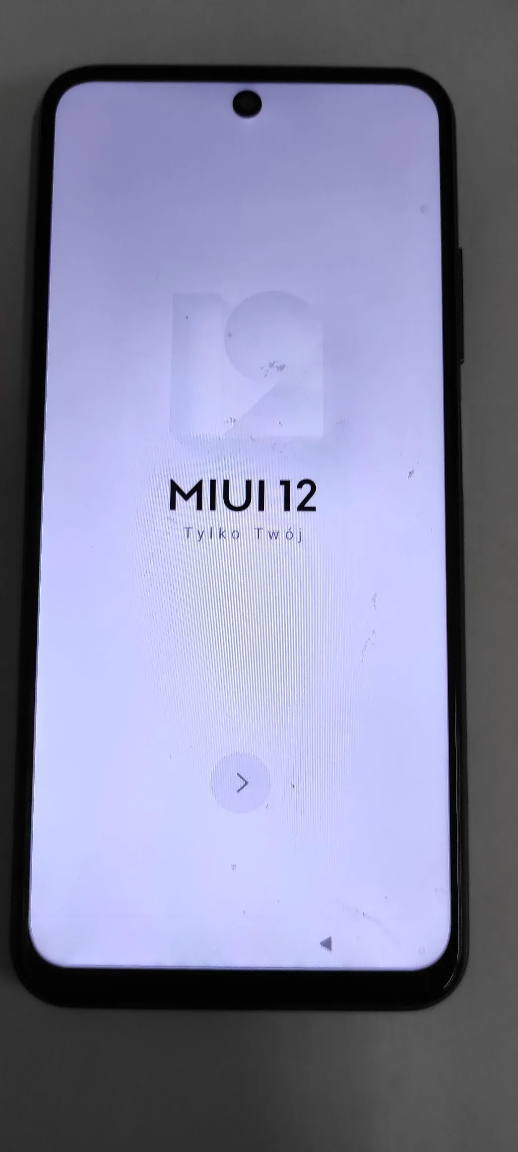 Smartfon z włączonym ekranem, na którym widnieje napis 'MIUI 12 Tylko Twój' oraz logo MIUI. Tekst i grafika są zorientowane pionowo względem obudowy telefonu, która leży poziomo. Na ekranie widoczny jest również przycisk strzałki skierowanej w górę oraz przednia kamera w formie otworu w lewym górnym rogu ekranu.