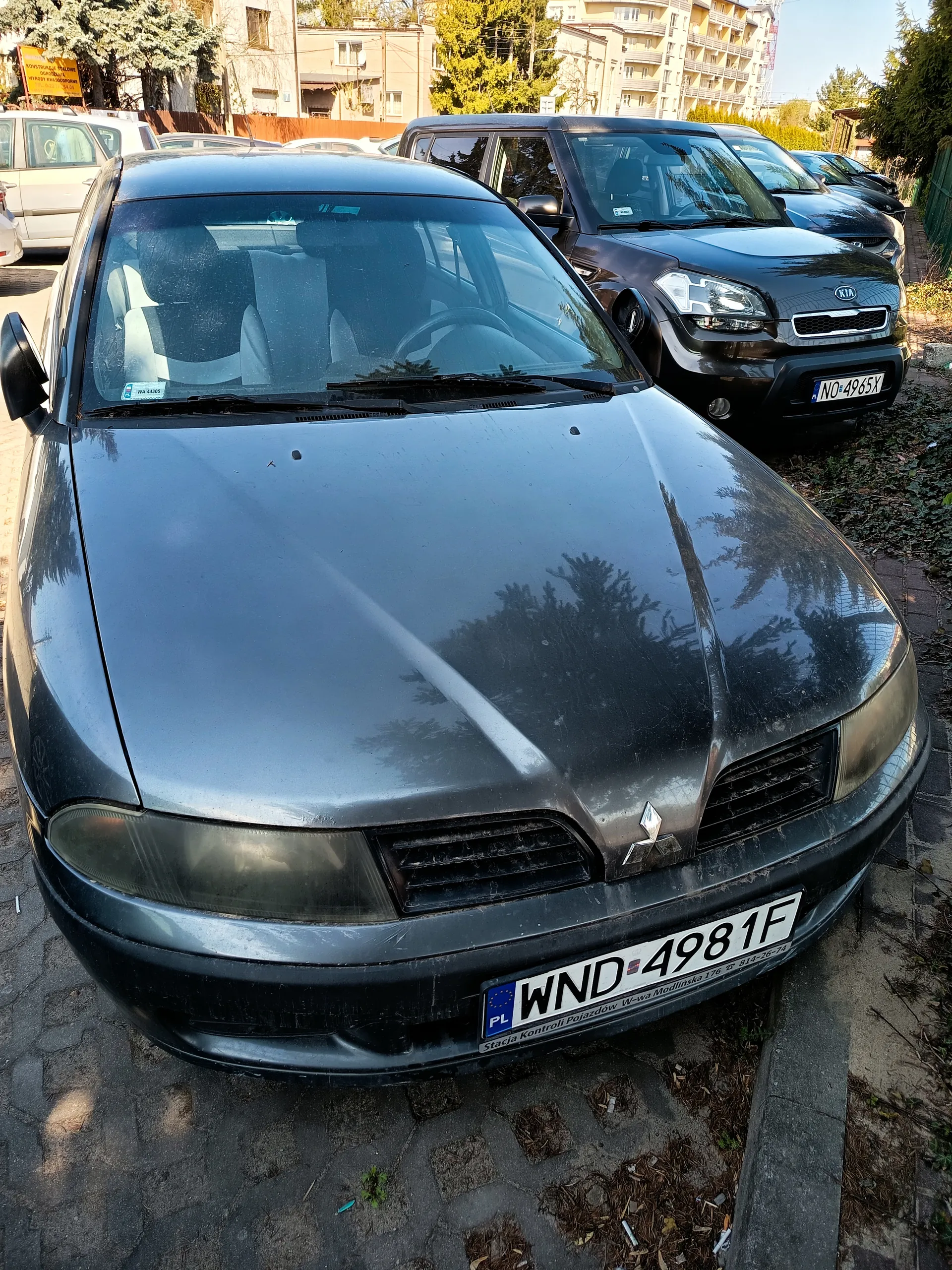 Przedni widok ciemnoszarego samochodu Mitsubishi z tablicą rejestracyjną WND 4981F (PL, Stacja Kontroli Pojazdów). W tle czarny samochód Kia z tablicą NO 4965X oraz inne zaparkowane pojazdy. Tło stanowią budynki mieszkalne, drzewa i brukowana powierzchnia z suchymi liśćmi. Światło słoneczne odbija się od maski samochodu.