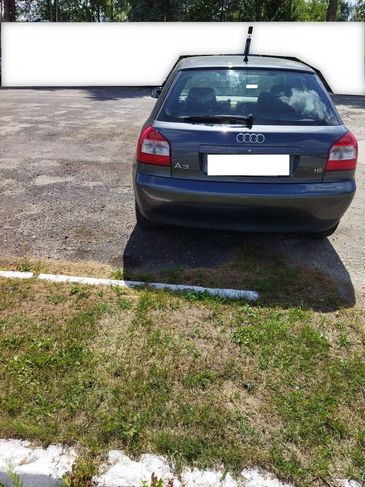 Widok z tyłu na ciemnoszary samochód marki Audi A3 zaparkowany na asfaltowym placu. Na klapie bagażnika widoczne jest logo Audi (cztery pierścienie), oznaczenie modelu 'A3' oraz oznaczenie silnika '1.6'. Tablica rejestracyjna jest zakryta białym prostokątem. W tle znajduje się biały mur i pas zielonych drzew. Na pierwszym planie widoczny jest fragment trawnika z białym krawężnikiem.