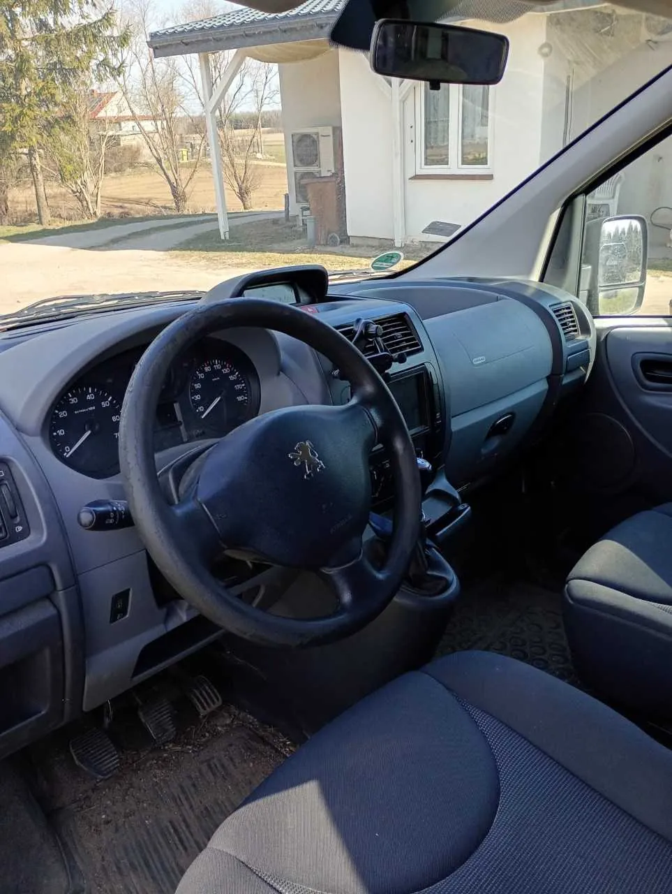 Wnętrze samochodu widziane z miejsca pasażera. Kierownica z logo Peugeot, deska rozdzielcza z licznikami prędkości i obrotomierzem, siedzenia w kolorze niebieskim z szarymi wstawkami. Przez przednią szybę widoczny biały dom z oknami, drzewa bez liści i droga ziemna. W tle dom z czerwonym dachem i sucha trawa.