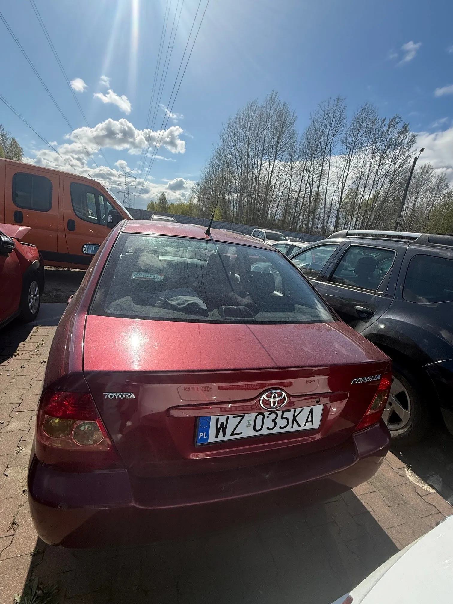 Czerwony samochód Toyota Corolla z tablicą rejestracyjną WZ 035KA (PL) zaparkowany na placu parkingowym. W tle widoczne są inne pojazdy, w tym pomarańczowy van i ciemny SUV, niebo z chmurami, drzewa oraz linie energetyczne. Słoneczny dzień.