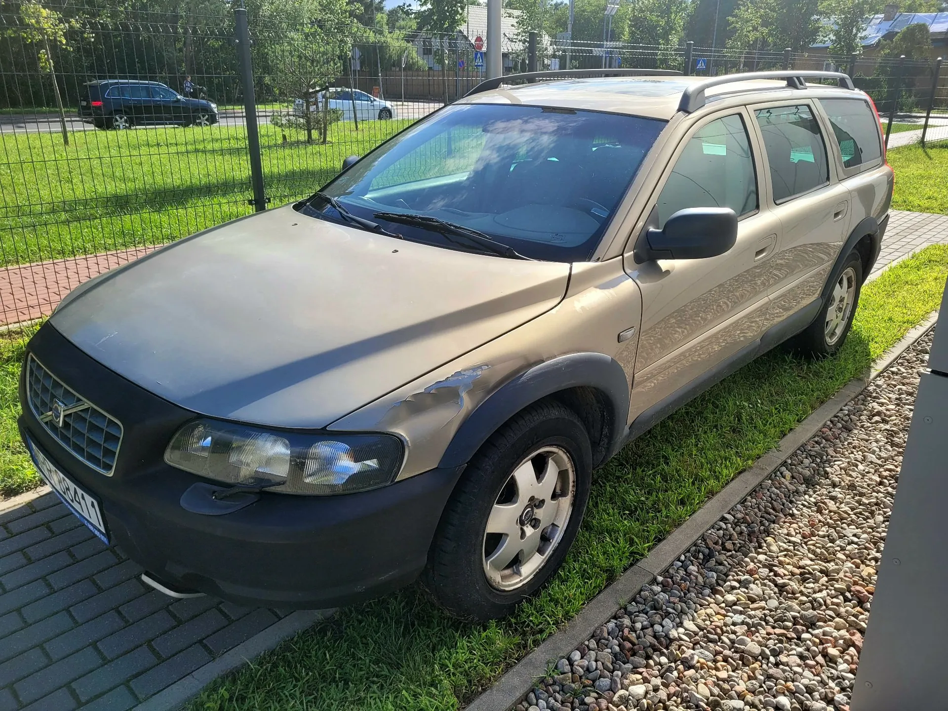 Złoty samochód marki Volvo w nadwoziu kombi, zaparkowany na trawie obok kostki brukowej i żwirowej ścieżki. Pojazd ma czarne plastikowe zderzaki i nadkola. W tle widoczny jest zielony trawnik, ogrodzenie z siatki oraz inne zaparkowane samochody i drzewa.