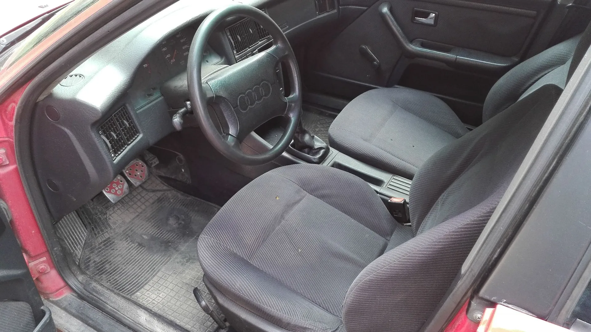 Wnętrze samochodu marki Audi z widokiem na stronę kierowcy. Widoczna jest czarna kierownica z logo czterech pierścieni, deska rozdzielcza z zegarami, dwa przednie fotele obite ciemnoszarym materiałem oraz gumowe dywaniki na podłodze. Drzwi kierowcy są otwarte.
