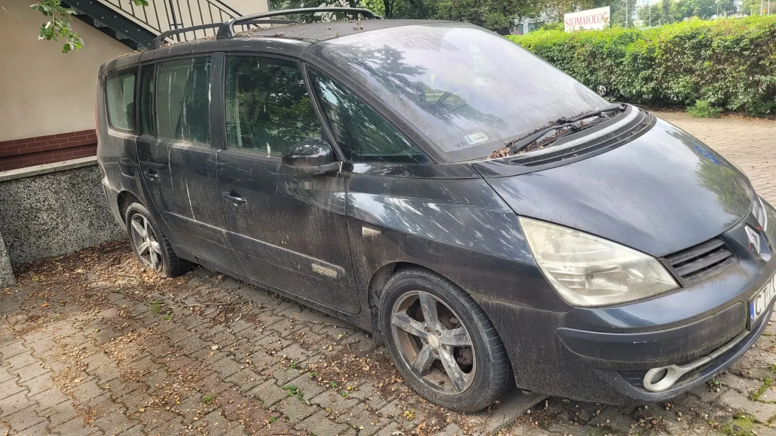 Brudny, ciemnogranatowy samochód typu minivan zaparkowany na kostce brukowej. Pojazd jest pokryty warstwą kurzu i błota, posiada relingi dachowe oraz pięcioramienne felgi aluminiowe. W tle widoczna jest zieleń krzewów oraz fragment budynku z napisem 'STOMATOLOG'.