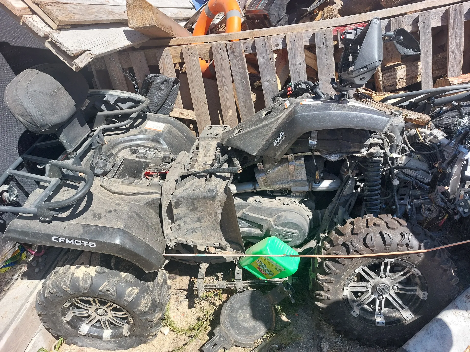 Czarny quad marki CFMOTO w stanie rozmontowanym, z odsłoniętym silnikiem i dużymi oponami pokrytymi błotem. Na boku widoczny napis 'CFMOTO'. W tle stos drewnianych desek i pomarańczowy pierścień ratunkowy. Przy quadzie zielony pojemnik z żółtą etykietą. Na ziemi widoczne fragmenty trawy i elementy mechaniczne.
