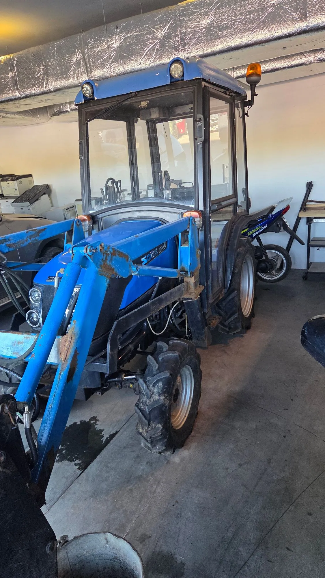 Niebieski traktor z kabiną i przednim ładowaczem, zaparkowany wewnątrz garażu lub warsztatu. Maszyna posiada duże, terenowe opony z wyraźnym bieżnikiem. Przedni ładowacz wykazuje ślady rdzy i zużycia. W tle widoczne są białe ściany, fragmenty sprzętów biurowych oraz srebrna izolacja termiczna na suficie.