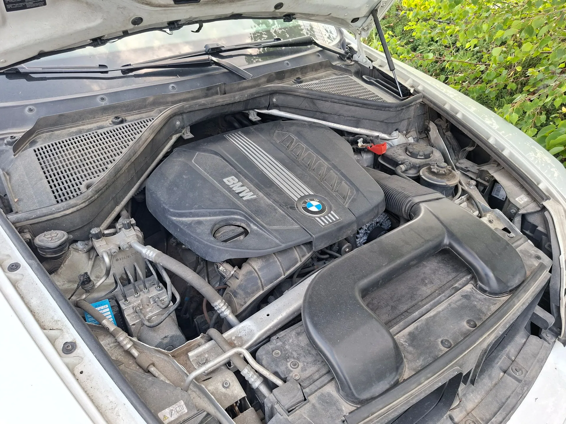 Widok z góry na komorę silnika białego samochodu marki BMW. W centrum znajduje się czarna plastikowa osłona silnika z białym napisem BMW oraz logo marki. Wokół widoczne są elementy mechaniczne, przewody, zbiorniki płynów oraz plastikowe obudowy. Maska samochodu jest otwarta i uniesiona.