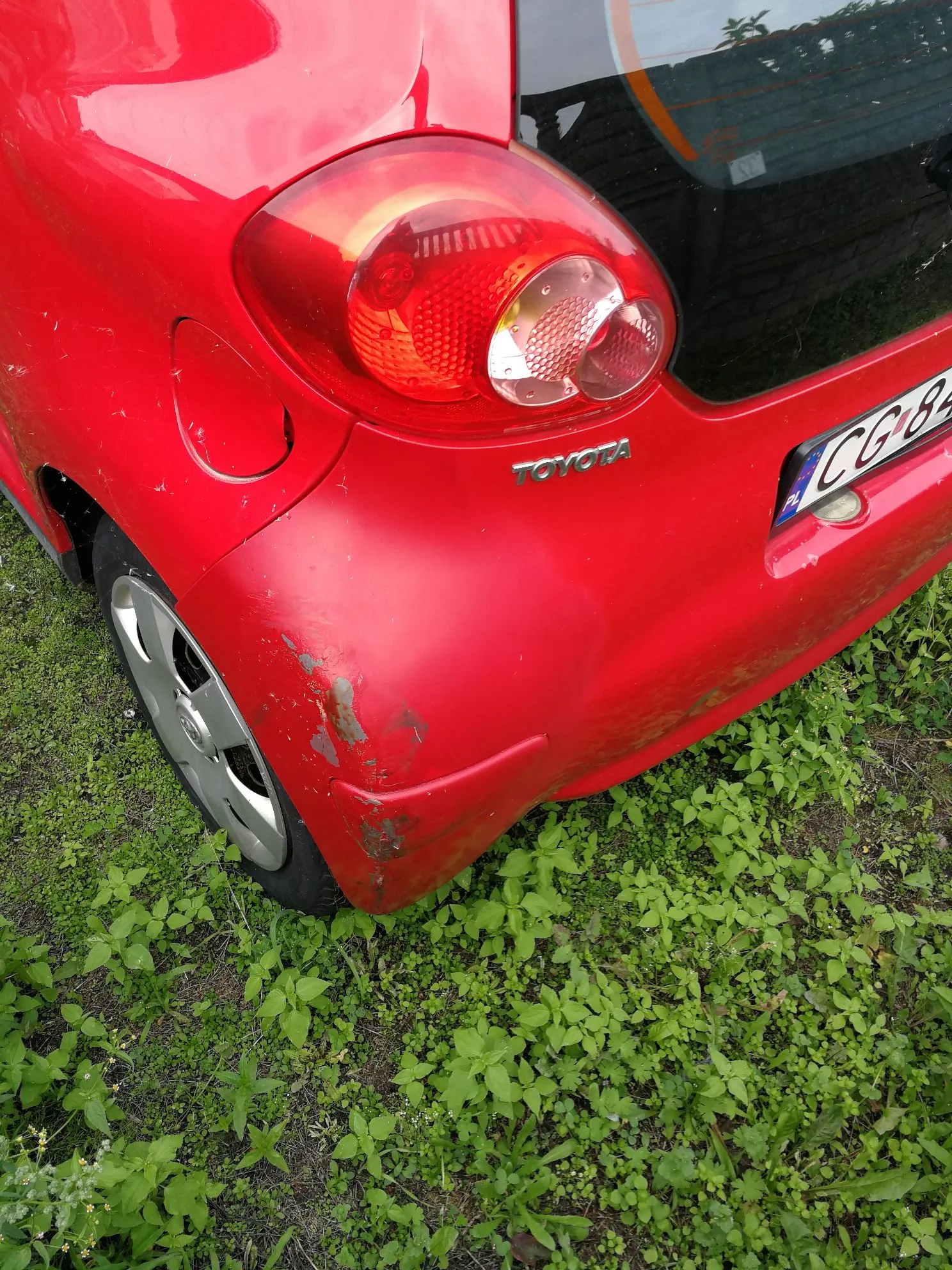 Zbliżenie na tylną, lewą część czerwonego samochodu marki Toyota. Widoczna jest tylna lampa, napis TOYOTA oraz fragment tablicy rejestracyjnej. Na tylnym zderzaku, nad kołem, znajdują się wyraźne uszkodzenia lakieru w postaci zadrapań i odprysków. Samochód stoi na trawie.