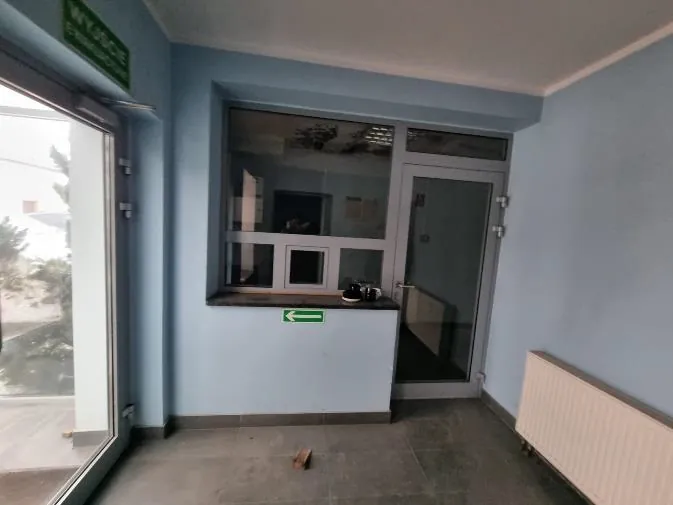 Wnętrze pomieszczenia z jasnoniebieskimi ścianami. Po lewej stronie duże okno z widokiem na zewnątrz, obok niego zielony znak. W centrum kontuar z oknem, na którym znajdują się przedmioty, poniżej zielona strzałka wskazująca w lewo. Po prawej stronie drzwi szklane i biały grzejnik. Podłoga z kafelkami, na niej mały przedmiot.