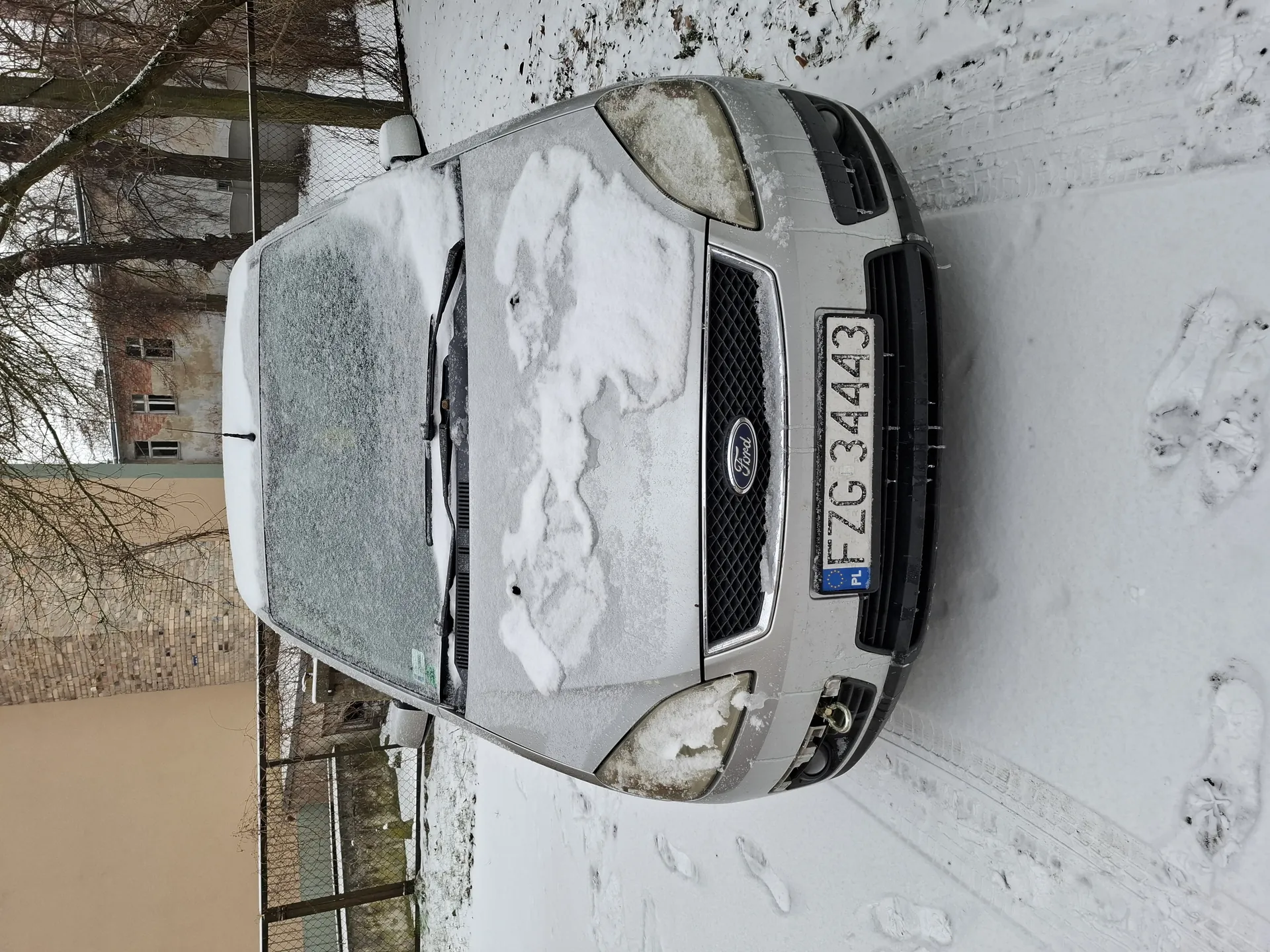 Srebrny samochód marki Ford pokryty śniegiem, widok z przodu. Na masce i przedniej szybie widoczne śnieżne odciski. Tablica rejestracyjna z białym tłem, niebieskim pasem z napisem PL i numerem FZG 34443. W tle budynki (beżowy i ceglany), metalowe ogrodzenie, drzewa bez liści oraz śnieżna powierzchnia z odciskami stóp i opon. Logo Ford na czarnej atrapie chłodnicy.