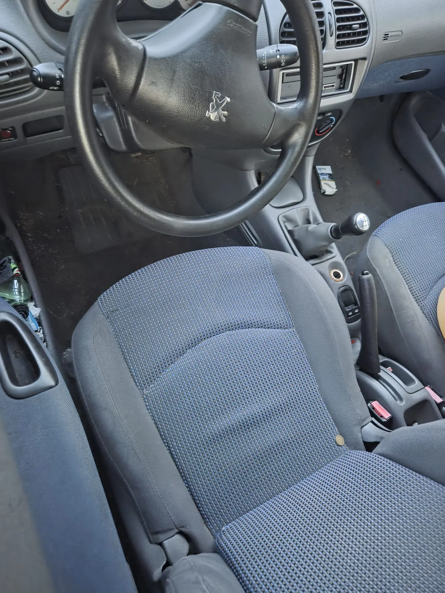 Wnętrze samochodu z widocznym kierownicą marki Peugeot, siedzeniami tapicerowanymi niebieską tkaniną, dźwignią zmiany biegów i hamulcem ręcznym. Na desce rozdzielczej widoczne są wentylacyjne otwory, przyciski oraz napis AIRBAG po prawej stronie. Na podłodze leżą różne przedmioty, w tym butelka i mały pakiet. Kierownica umieszczona jest centralnie, a siedzenia zajmują przestrzeń przednią.
