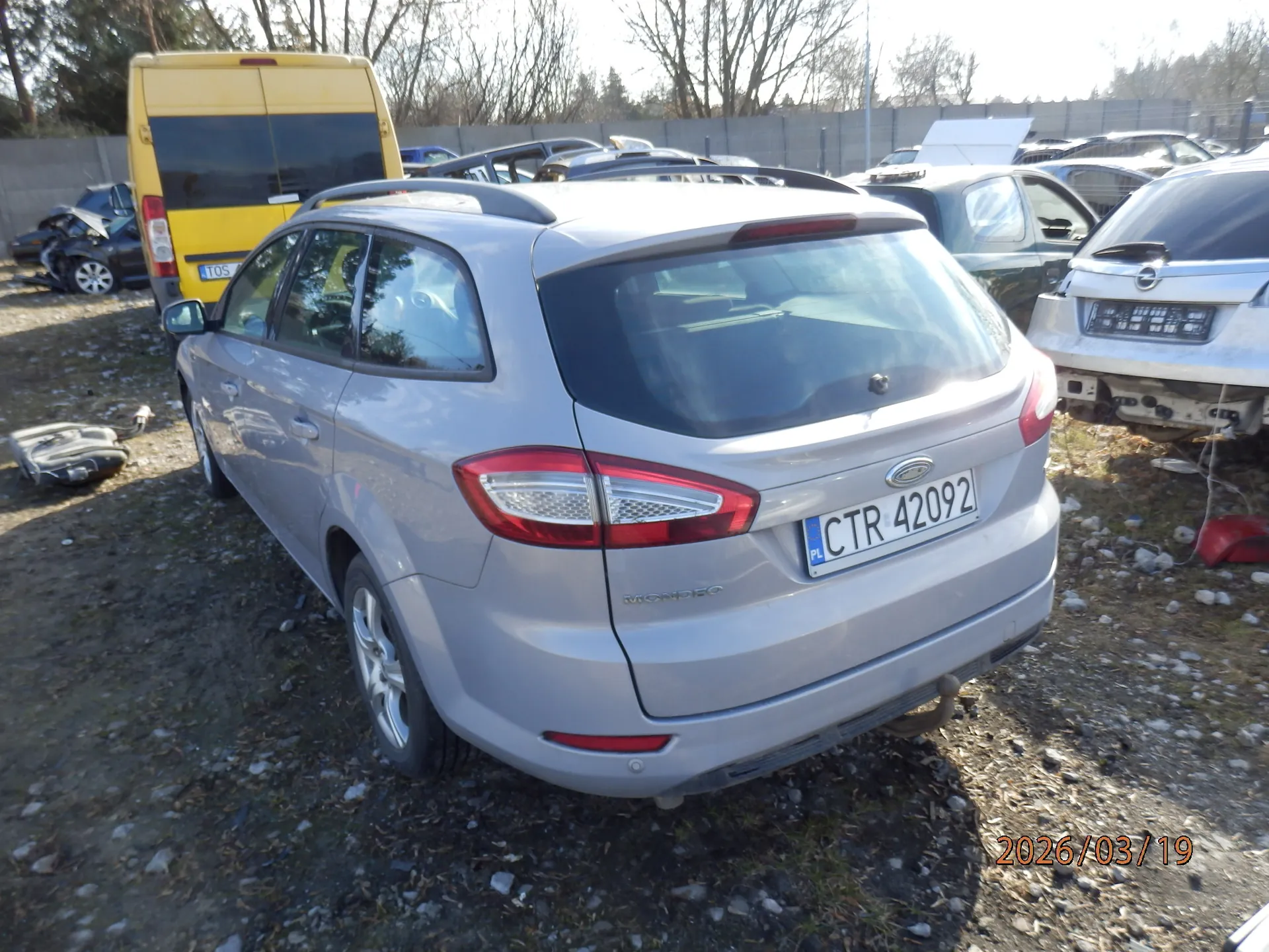 Srebrny samochód osobowy typu kombi marki Ford Mondeo z tablicą rejestracyjną CTR 42092 (PL) na tle stacji złomowania. W tle widoczne są inne uszkodzone pojazdy, w tym żółty van z tablicą T05, otoczone ogrodzeniem i drzewami bez liści. Na ziemi żwir i fragmenty części samochodowych. W prawym dolnym rogu obrazu widoczna data 2026/03/19.