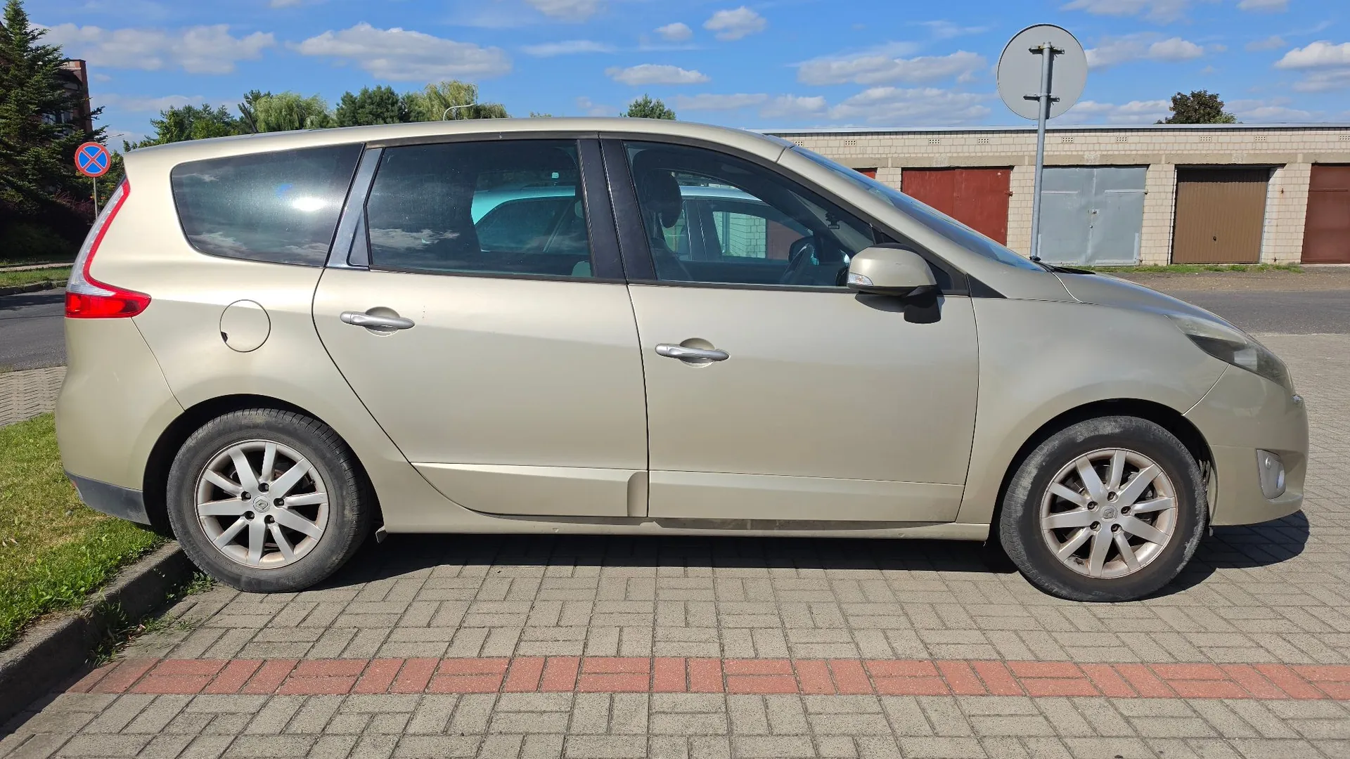 Profil boczny beżowego samochodu typu minivan zaparkowanego na kostce brukowej. W tle widoczne są garaże z brązowymi i szarymi drzwiami, niebo z lekkimi chmurami oraz znak drogowy.
