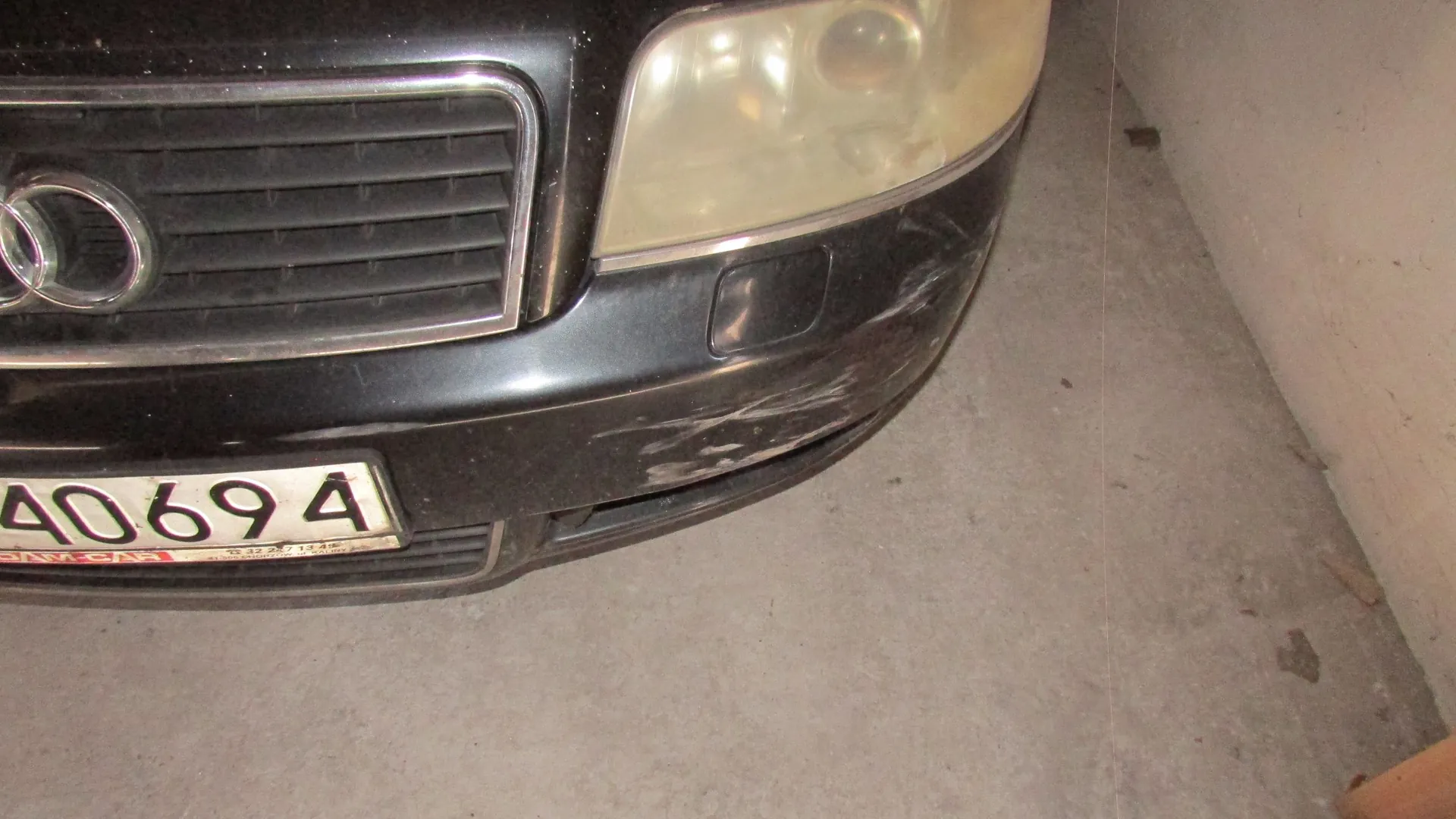 Zbliżenie na przednią część czarnego samochodu marki Audi. Widoczny jest fragment grilla z logo, zmatowiony reflektor oraz przedni zderzak z widocznymi zarysowaniami i przetarciami lakieru po prawej stronie. W dolnej części widoczny jest fragment tablicy rejestracyjnej z numerami 40694.