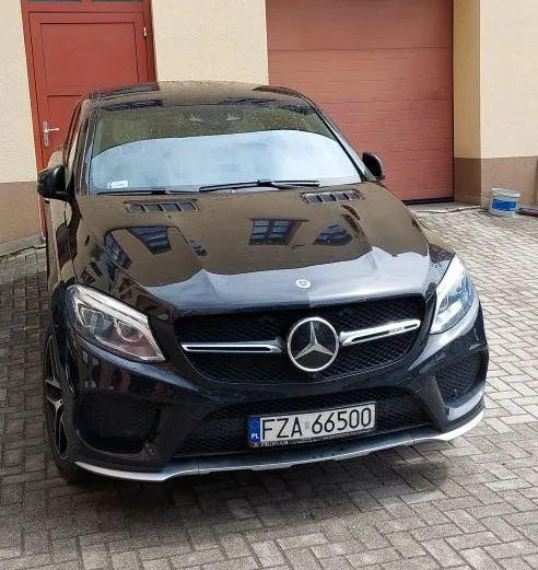 Czarny samochód Mercedes-Benz SUV zaparkowany na brukowanej powierzchni przed budynkiem. Widoczna przód pojazdu z logo Mercedes-Benz na atrapie chłodnicy oraz tablicą rejestracyjną FZA 66500. W tle czerwone drzwi i brązowa brama garażowa. Budynek posiada beżowe ściany i niebiesko-białą pojemnik na prawo od bramy.