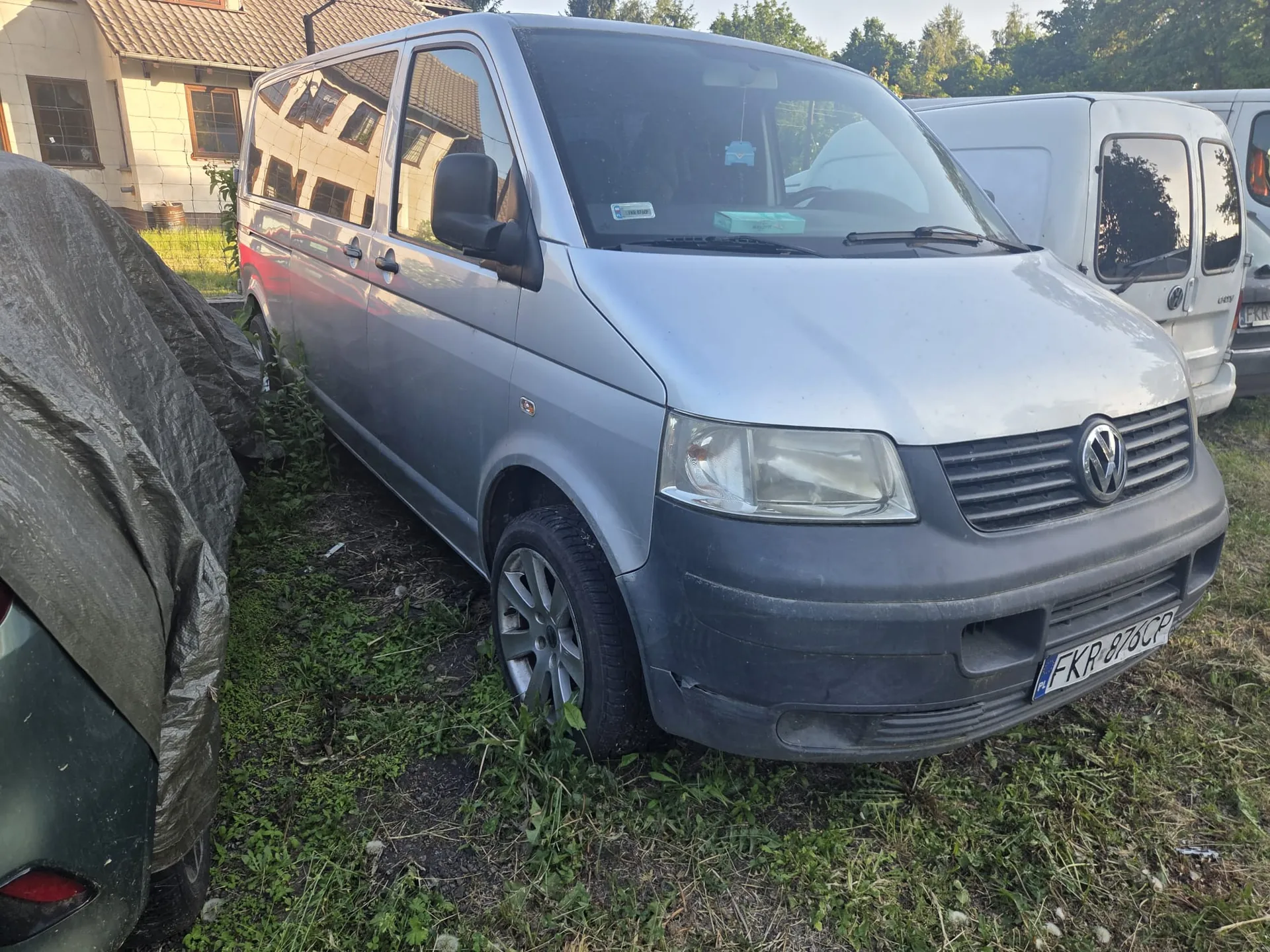 Srebrny samochód dostawczy Volkswagen z tablicą rejestracyjną FKR 876CP zaparkowany na trawie. W tle biały samochód dostawczy, dom z dachem krytym dachówką i drzewa. Po lewej stronie częściowo zakryty czarną folią pojazd. Trawa wokół jest nieco porośnięta, a na przednim zderzaku i kołach widoczny brud. W tylnej szybie srebrnego Volkswagena widoczny niebieski przedmiot.