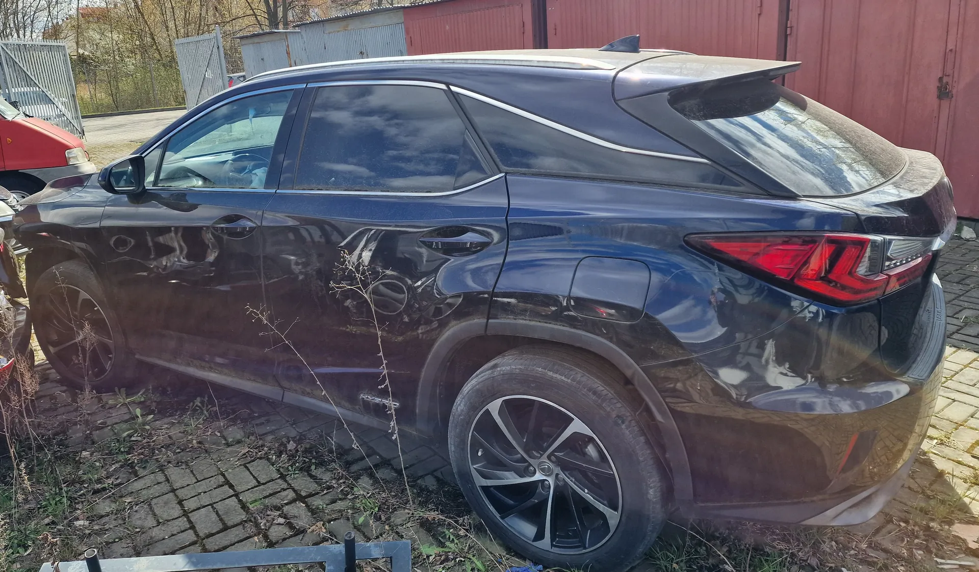 Ciemnoniebieski SUV zaparkowany na brukowanej powierzchni. Widok z boku i tyłu, z widocznymi czerwonymi światłami tylnymi, czarnymi felgami i lusterkiem bocznym. W tle czerwony budynek, metalowe ogrodzenie, fragment czerwonego samochodu po lewej stronie oraz suche rośliny i trawa na ziemi. Na pierwszym planie metalowa konstrukcja.