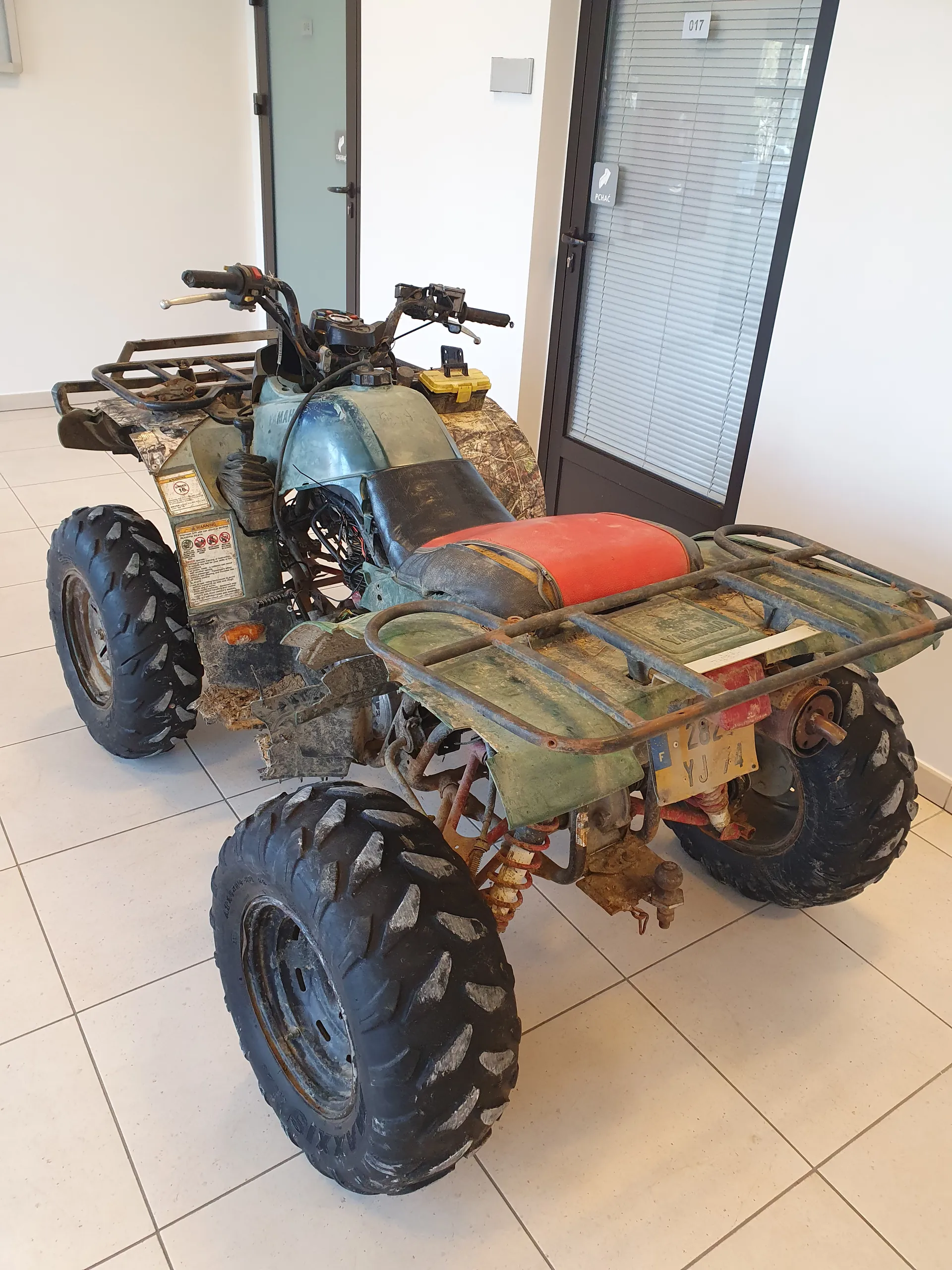 Zdjęcie przedstawia zielony quad z czerwonym siedzeniem, pokryty rdzą i brudem, stojący na białej płytkowej podłodze w pomieszczeniu. Quad ma duże czarne opony z widocznym wzorem, żółty tabliczki rejestracyjnej z napisem YJ 282 oraz naklejki ostrzegawcze na kadłubie. W tle widać szklane drzwi z napisem PUSH i oznaczeniem 017 oraz okno z roletą. Pojazd znajduje się w pozycji nieprawidłowo zorientowanej względem horyzontu.