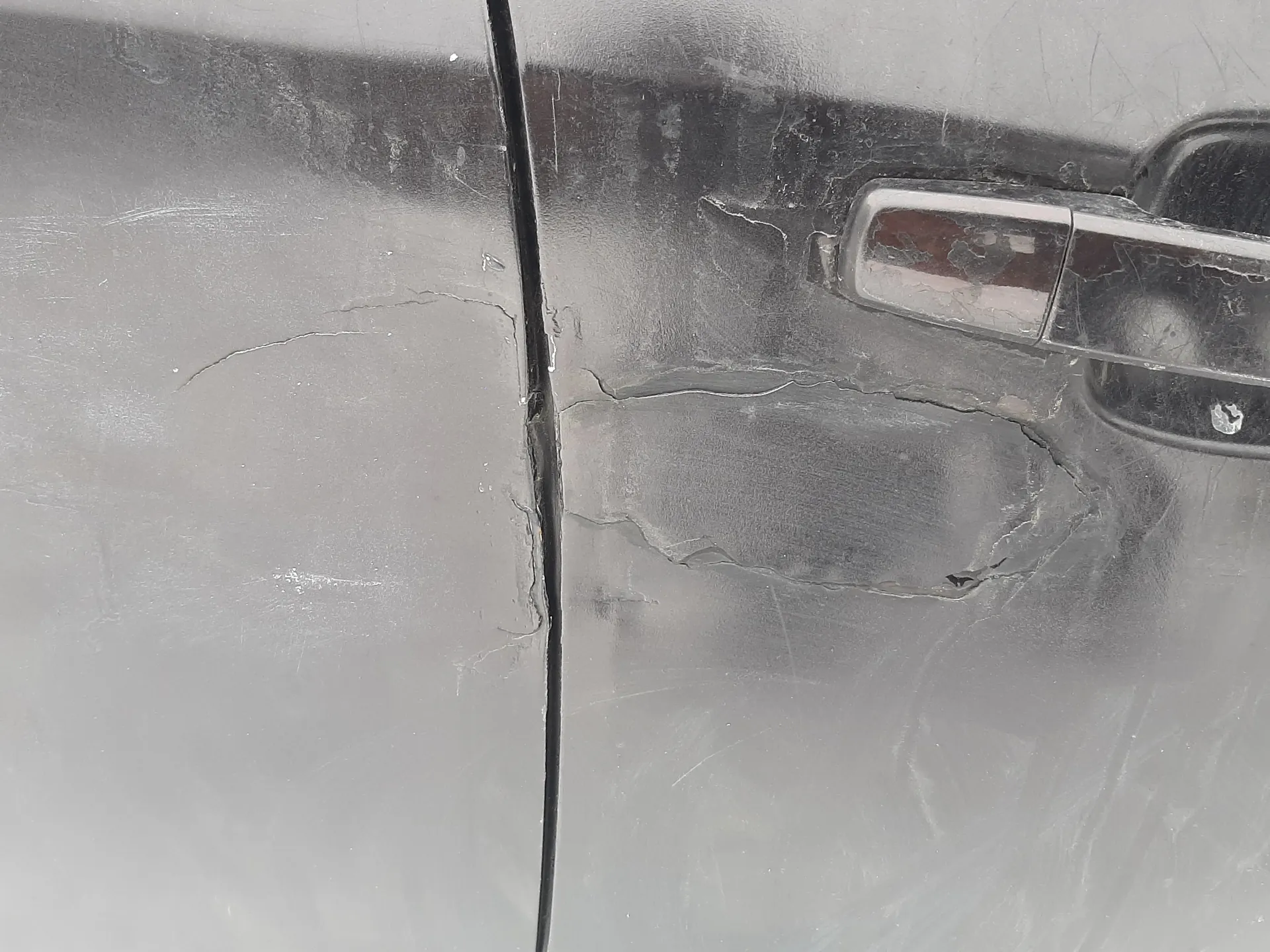 Zdjęcie przedstawia uszkodzoną czarną karoserię samochodu. Widoczna jest duża wgnieciona plama z pęknięciami lakieru wokół klamki drzwi. Na powierzchni znajdują się liczne zadrapania i ślady uszkodzeń mechanicznych. Klamka drzwi jest metalowa, częściowo zarysowana.